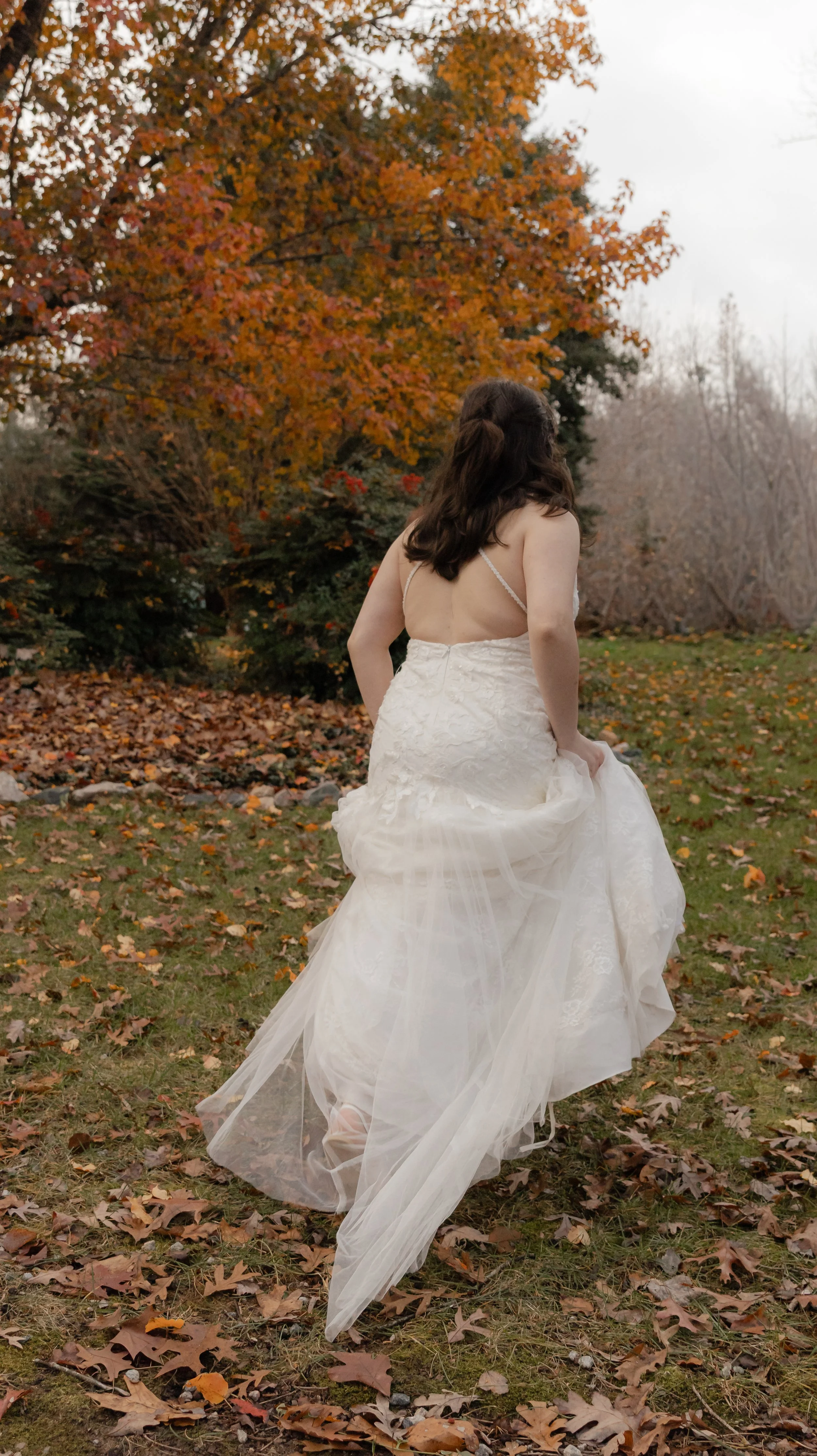 A woman in a white wedding dress walks away in an autumn outdoor setting with fallen leaves and trees with orange foliage.