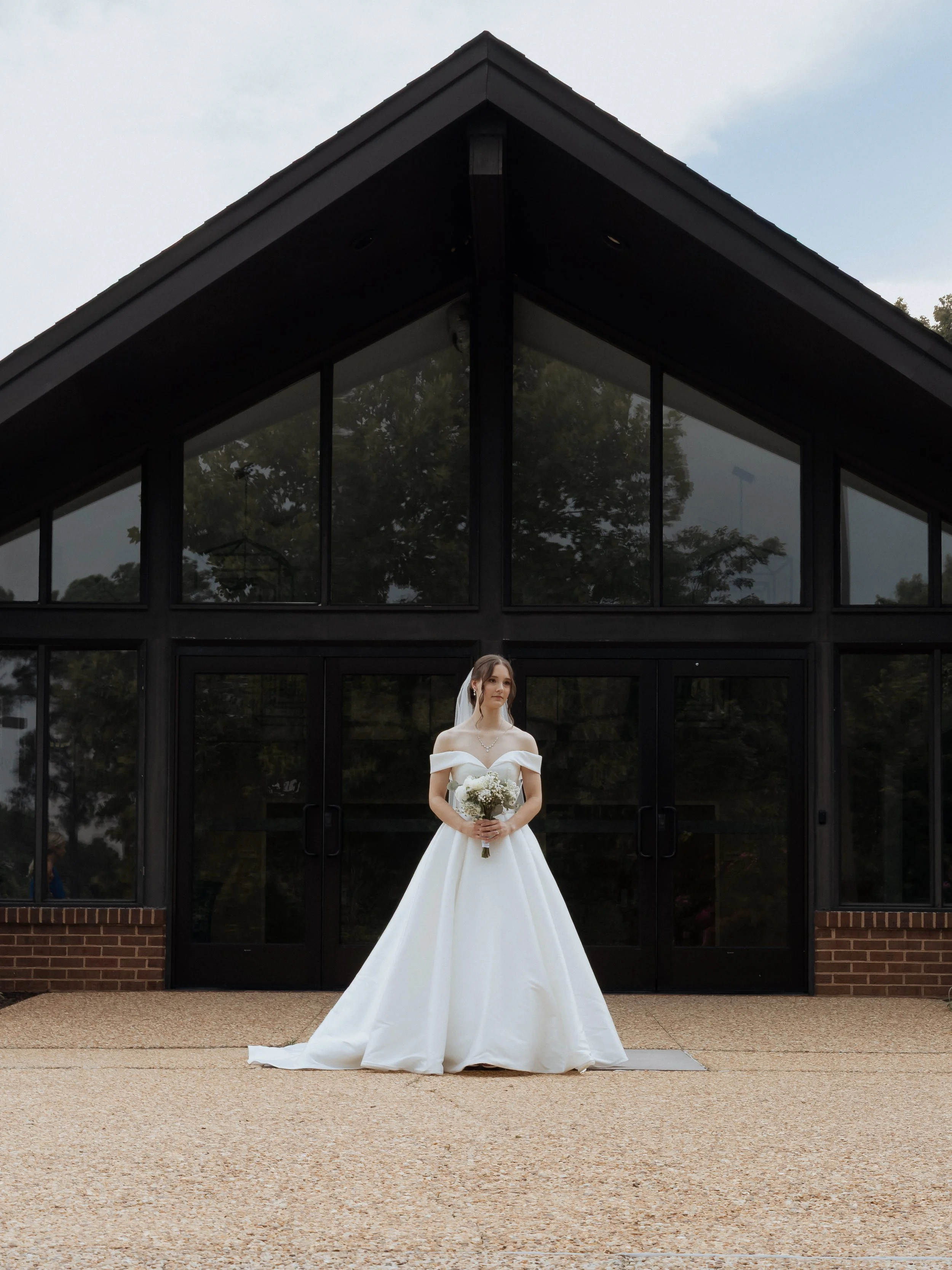 A bride holding a bouquet standing in front of a building with large windows wearing a white wedding gown and a veil.