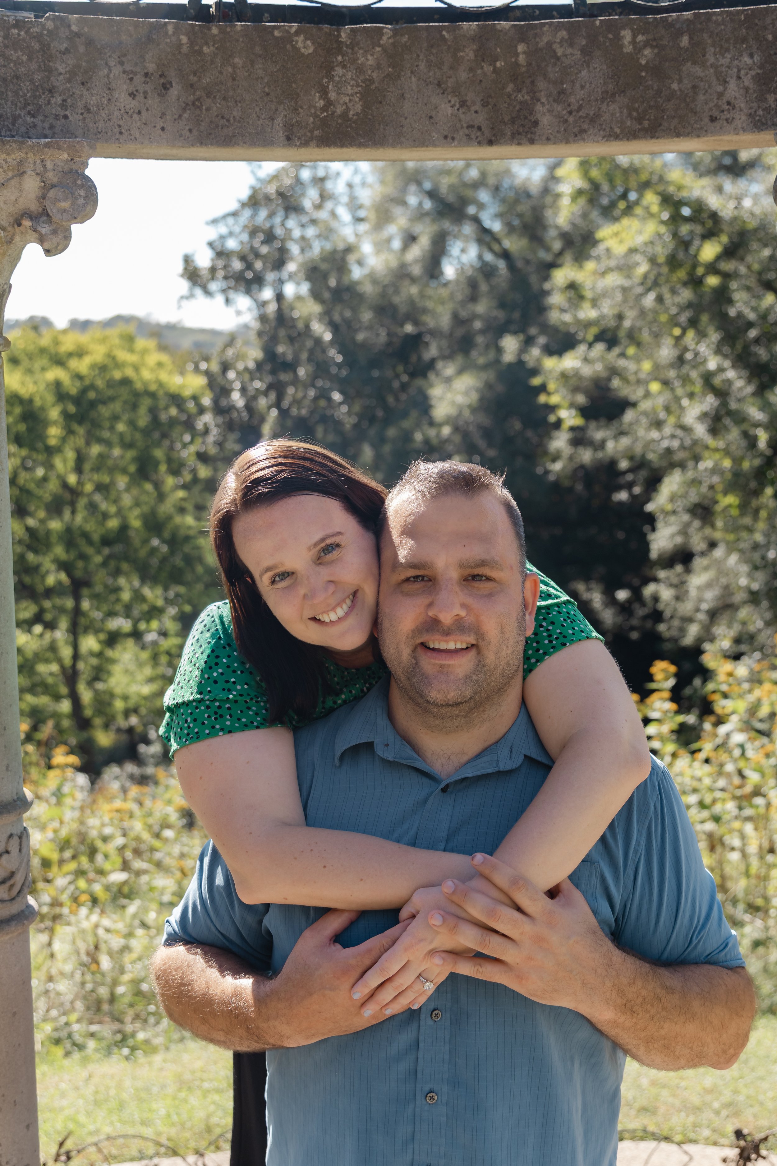 A couple enjoying a sunny day outdoors, with the woman hugging from behind while resting her head on the man's shoulder. They are smiling, and the background features green trees and a partly cloudy sky.