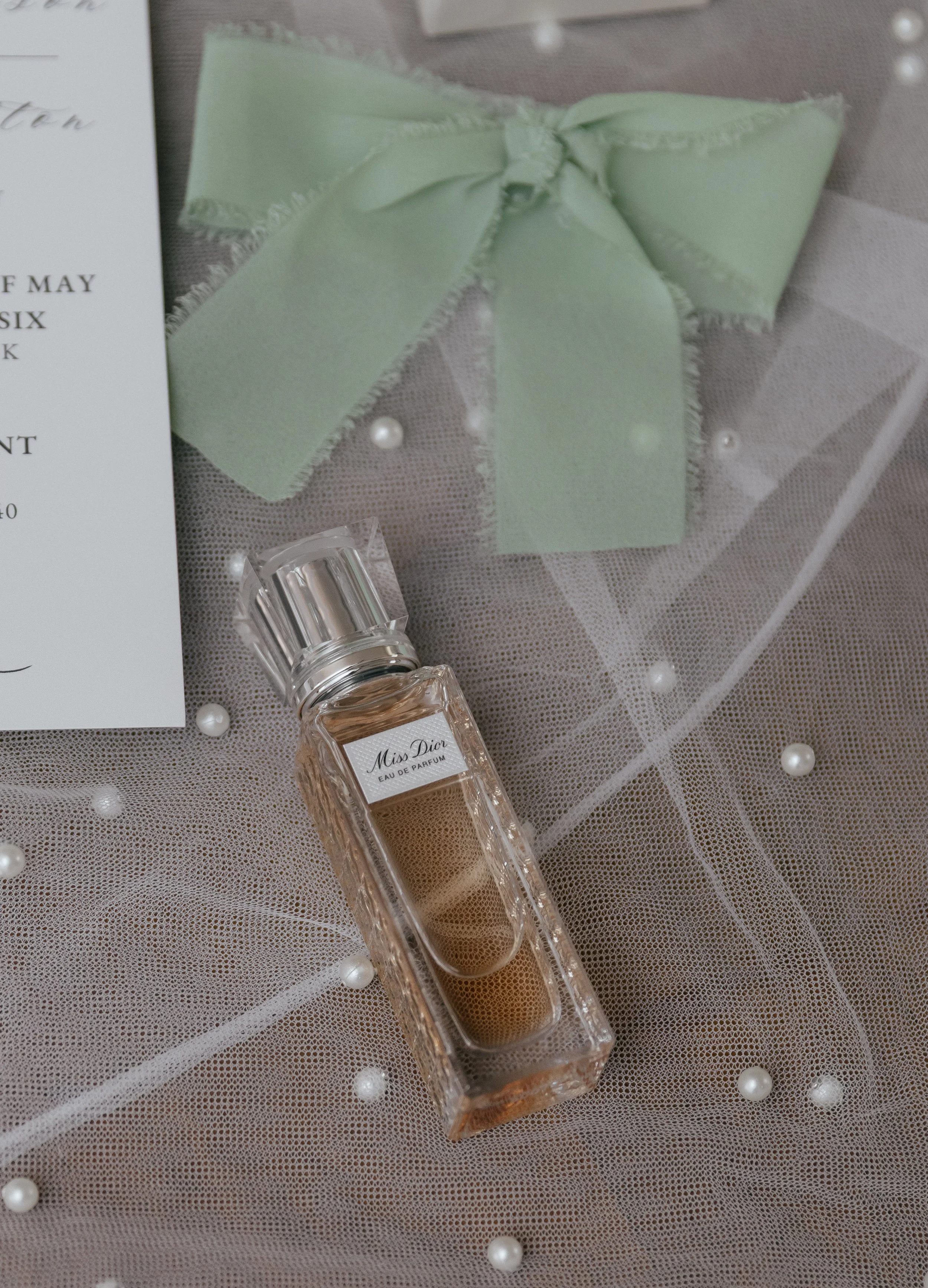 A bottle of Miss Dior Eau de Parfum on a mesh surface, with a green ribbon bow and a partially visible invitation or card nearby.