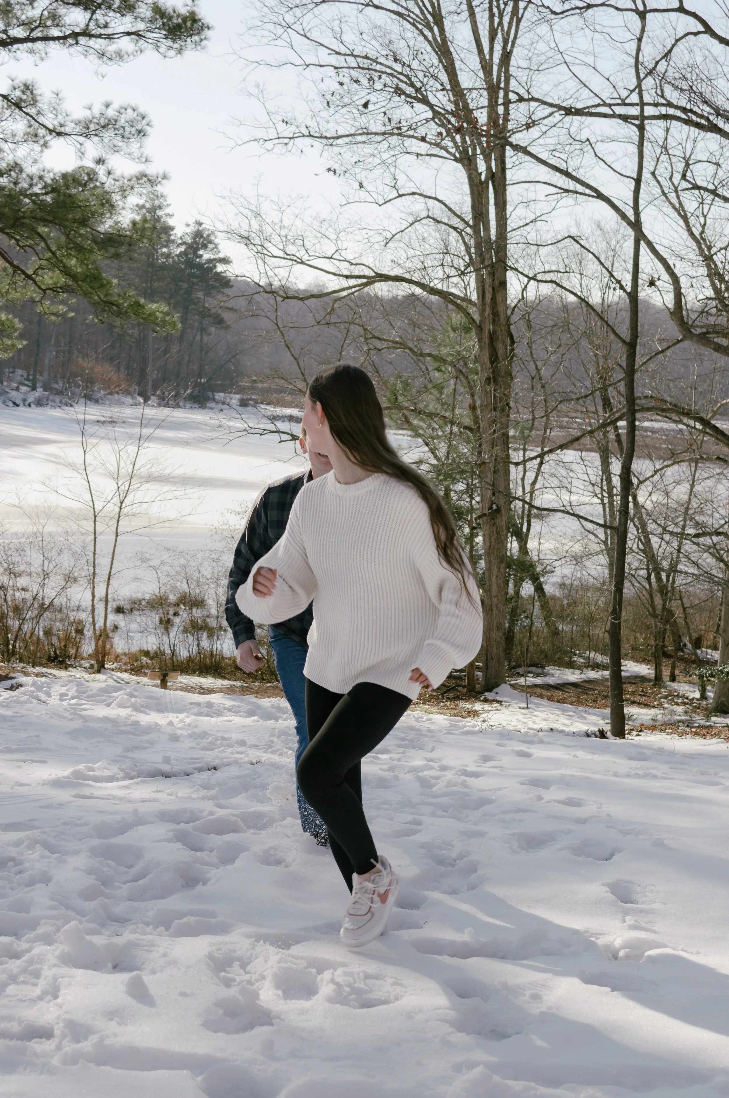 A couple in winter clothing walking through snow in a park with trees and a frozen lake in the background.