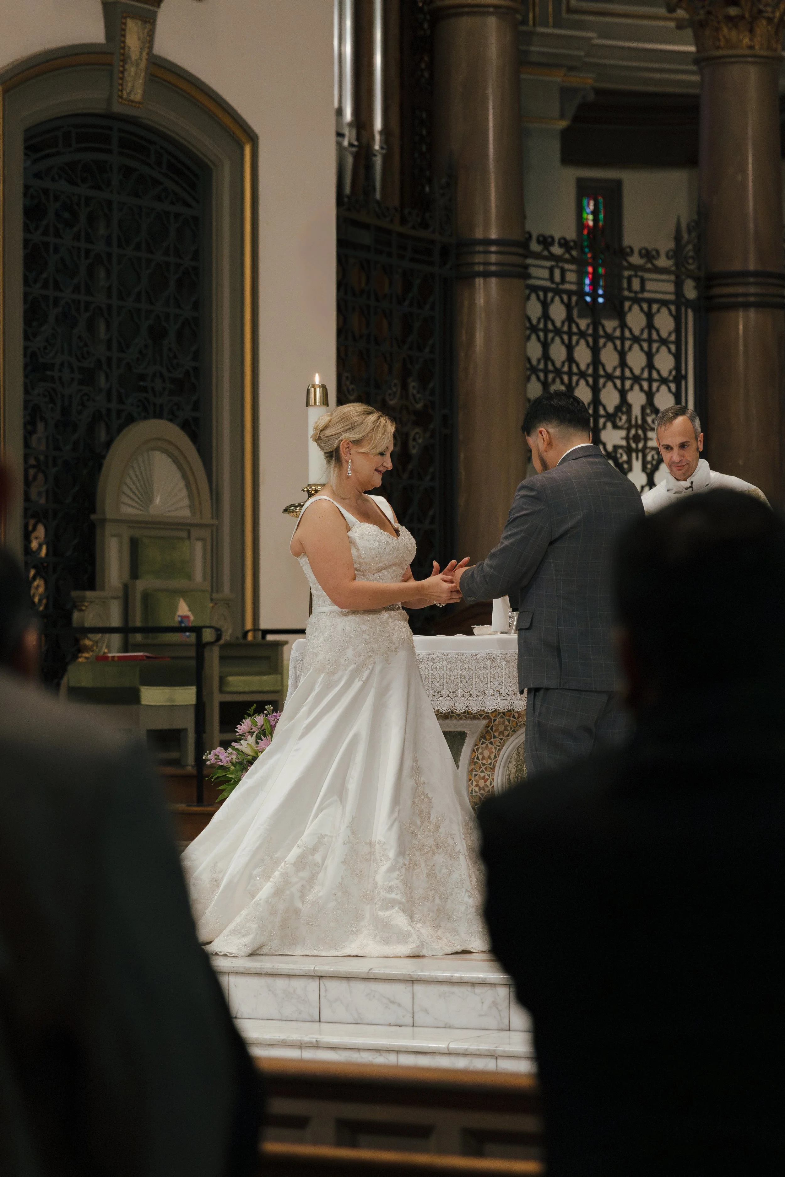 A bride and groom exchanging vows during a wedding ceremony inside a church with ornate ironwork and stained glass windows, with a priest or officiant in the background.