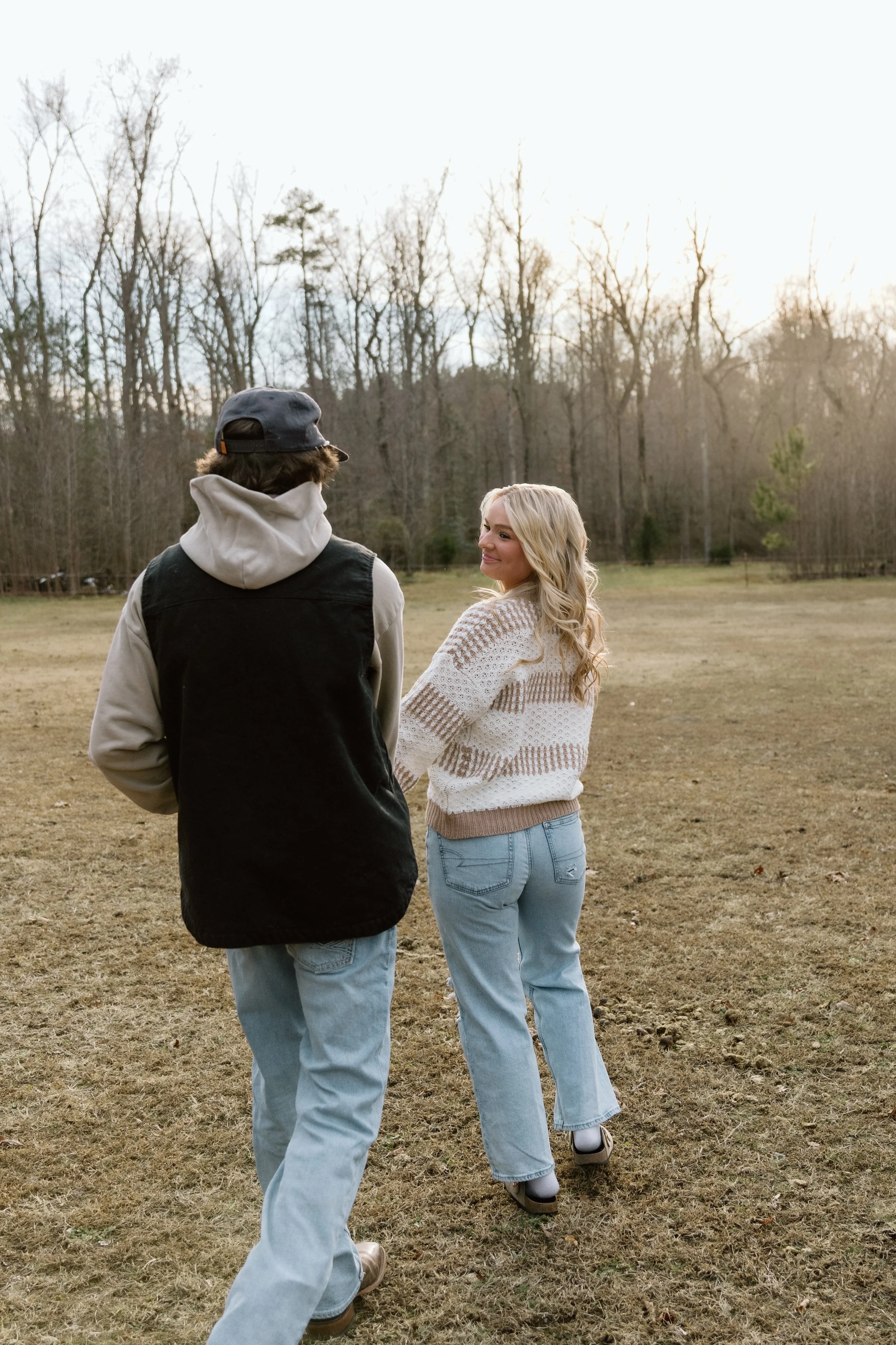 A young woman with long blonde hair smiling at a young man with brown hair, both walking outdoors on a grassy field with trees in the background during late afternoon or early evening.