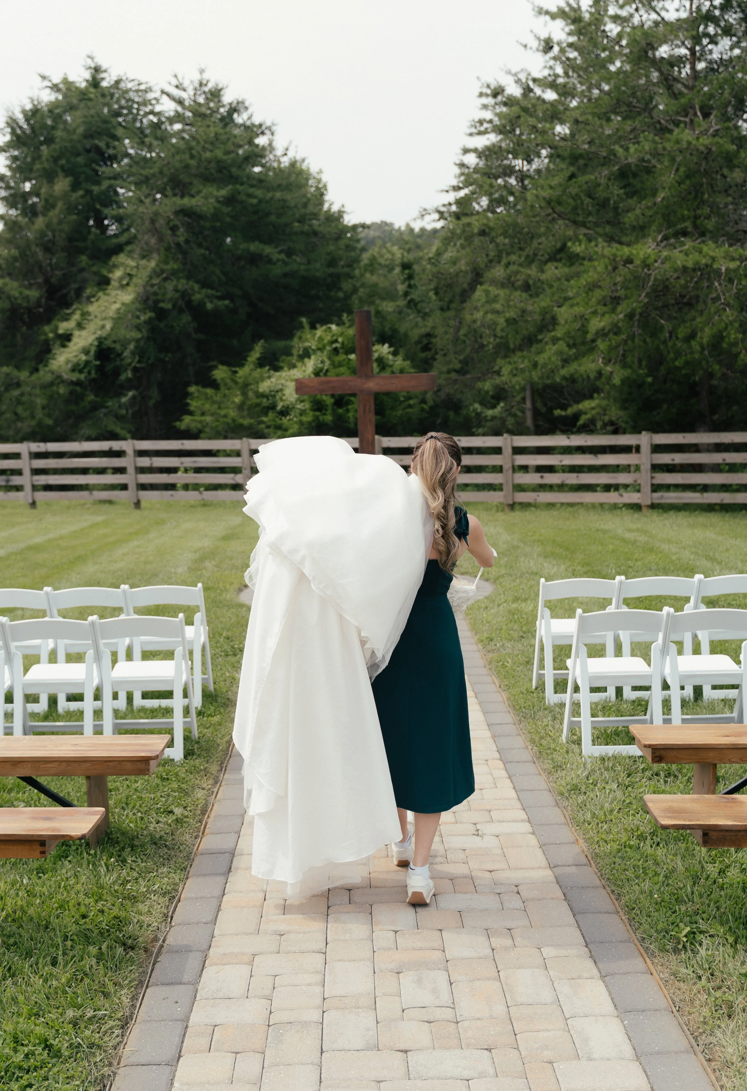 A woman in a green dress and white sneakers carrying a wedding dress down the aisle of a wedding venue in Virginia, walking towards a cross at the end of the aisle.