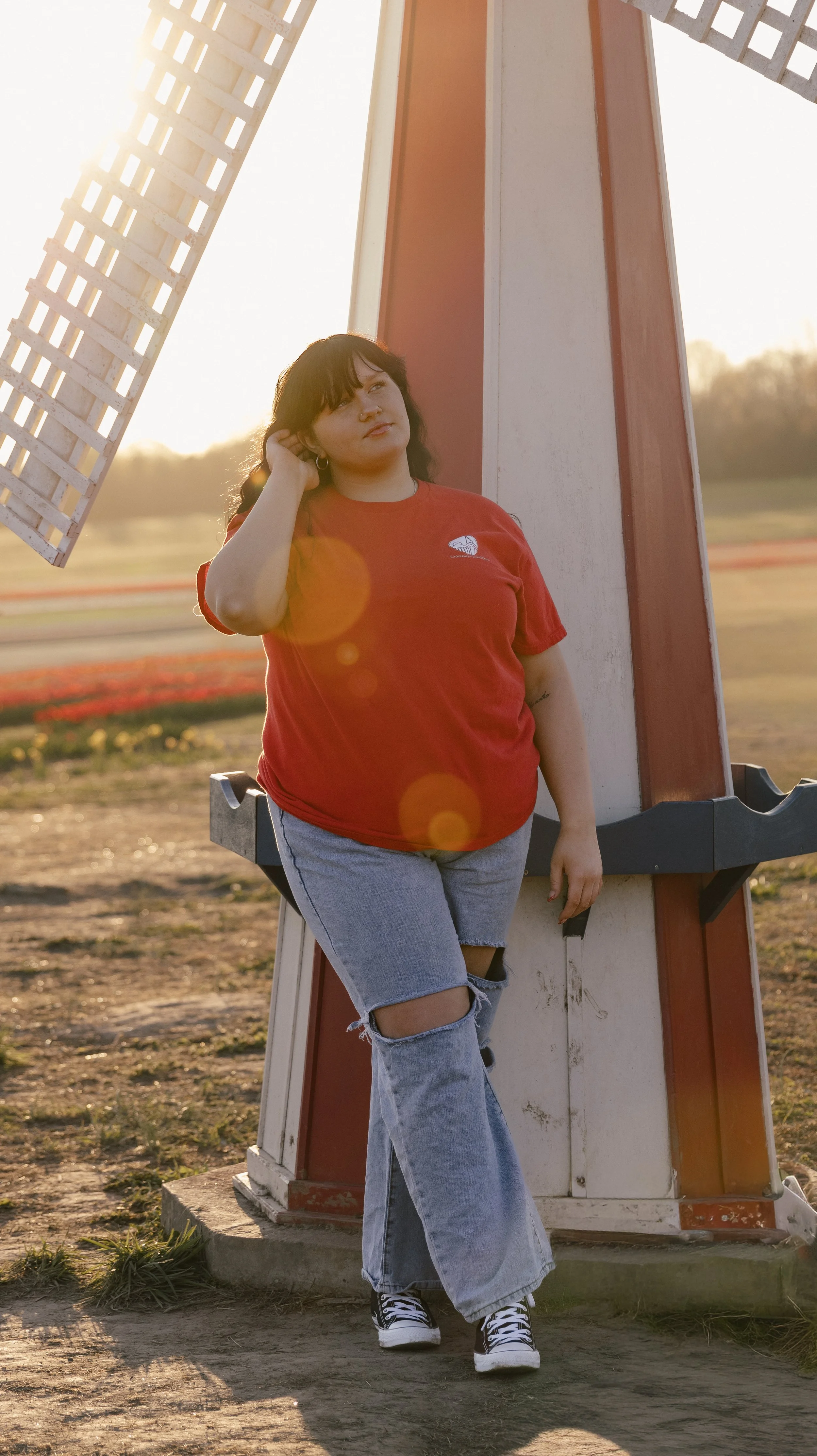 A woman with dark hair wearing a red T-shirt and ripped jeans standing next to a windmill at sunset.