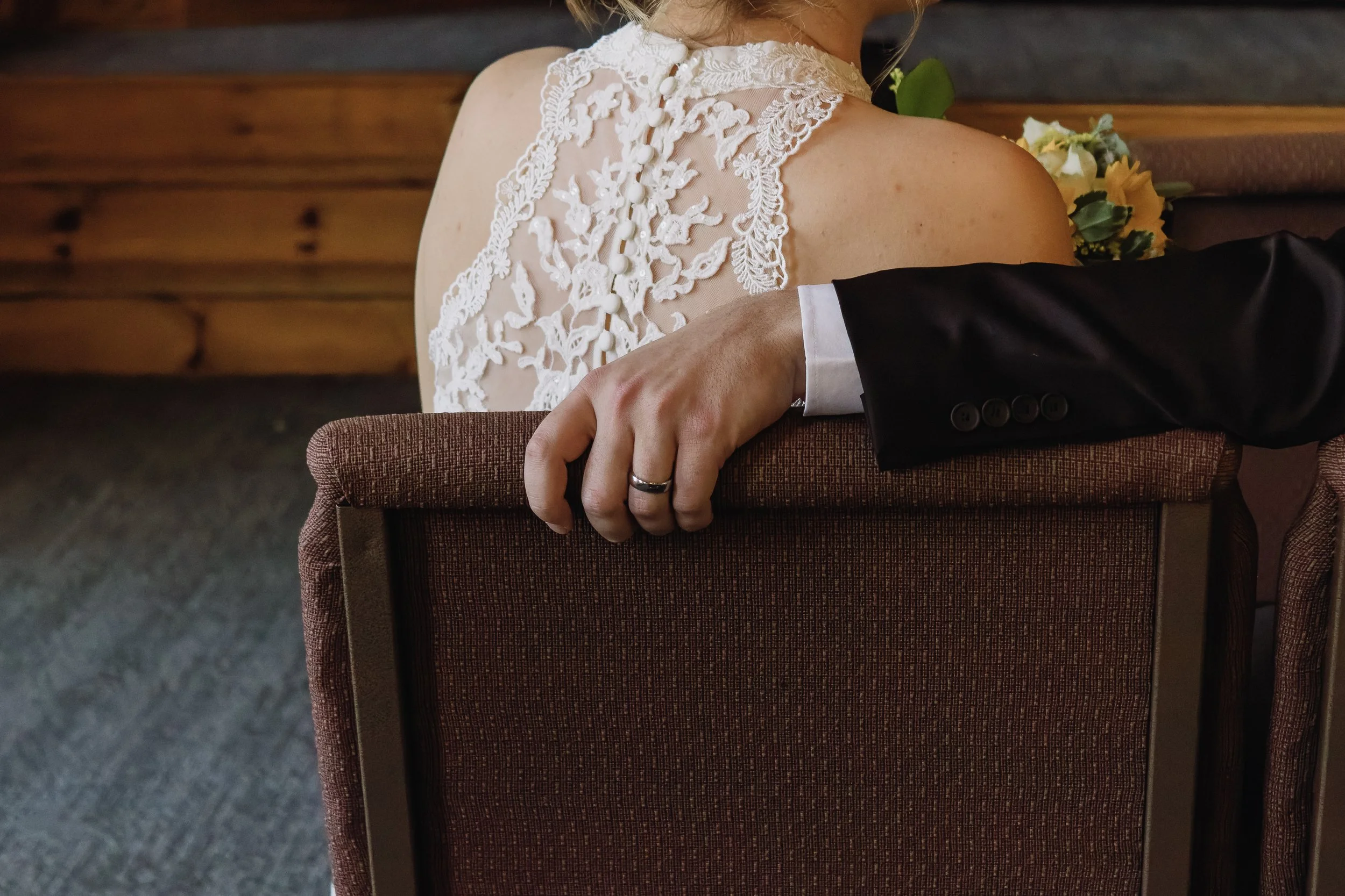 Close-up of a couple sitting together, with the man's hand wearing a wedding band resting on the back of a chair behind the woman, who is wearing a lace wedding dress.
