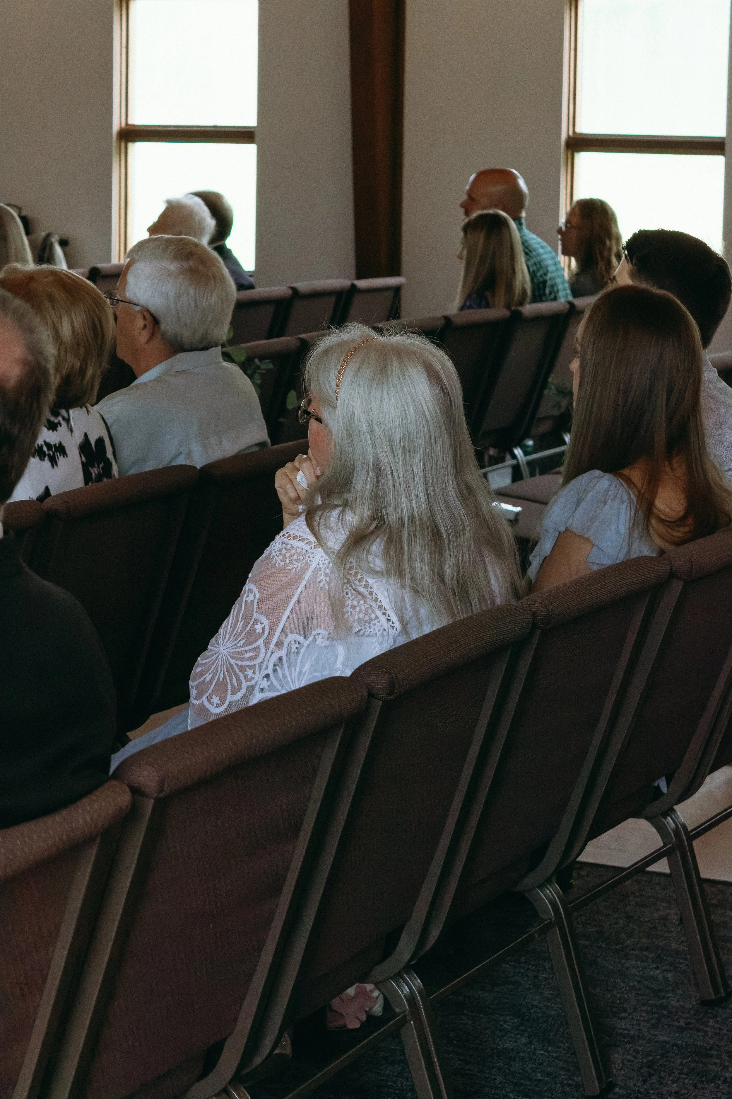 People sitting and listening in a church or auditorium, facing forward, with windows in the background.