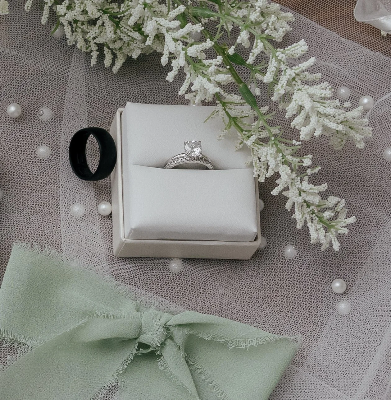 A white jewelry box with a diamond ring inside, surrounded by white flowers, a black ring, and a pale green bow on a textured fabric with pearl-like beads.