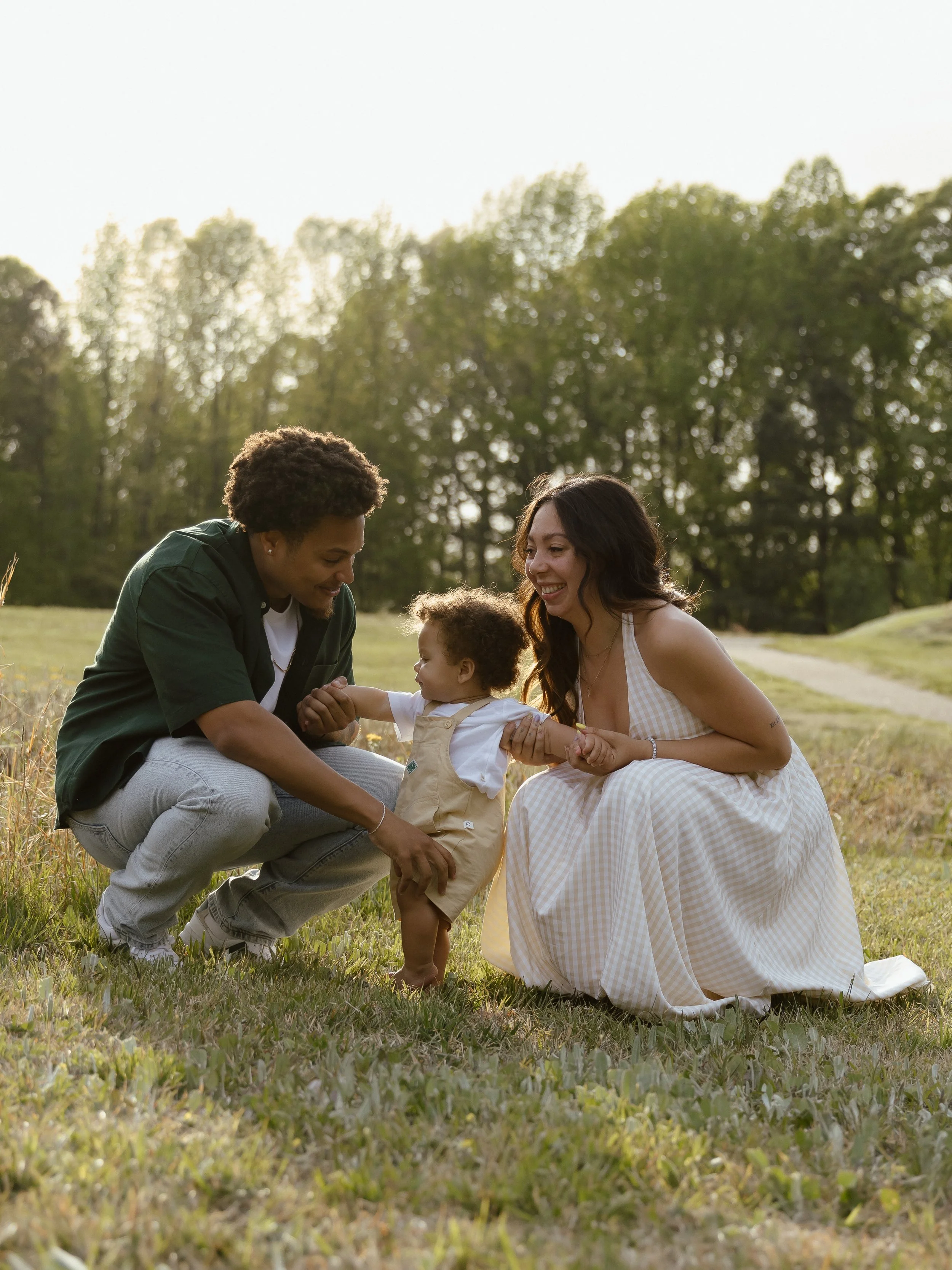 Family of three playing together outdoors in a grassy field with trees in the background, a young man and woman with a toddler.