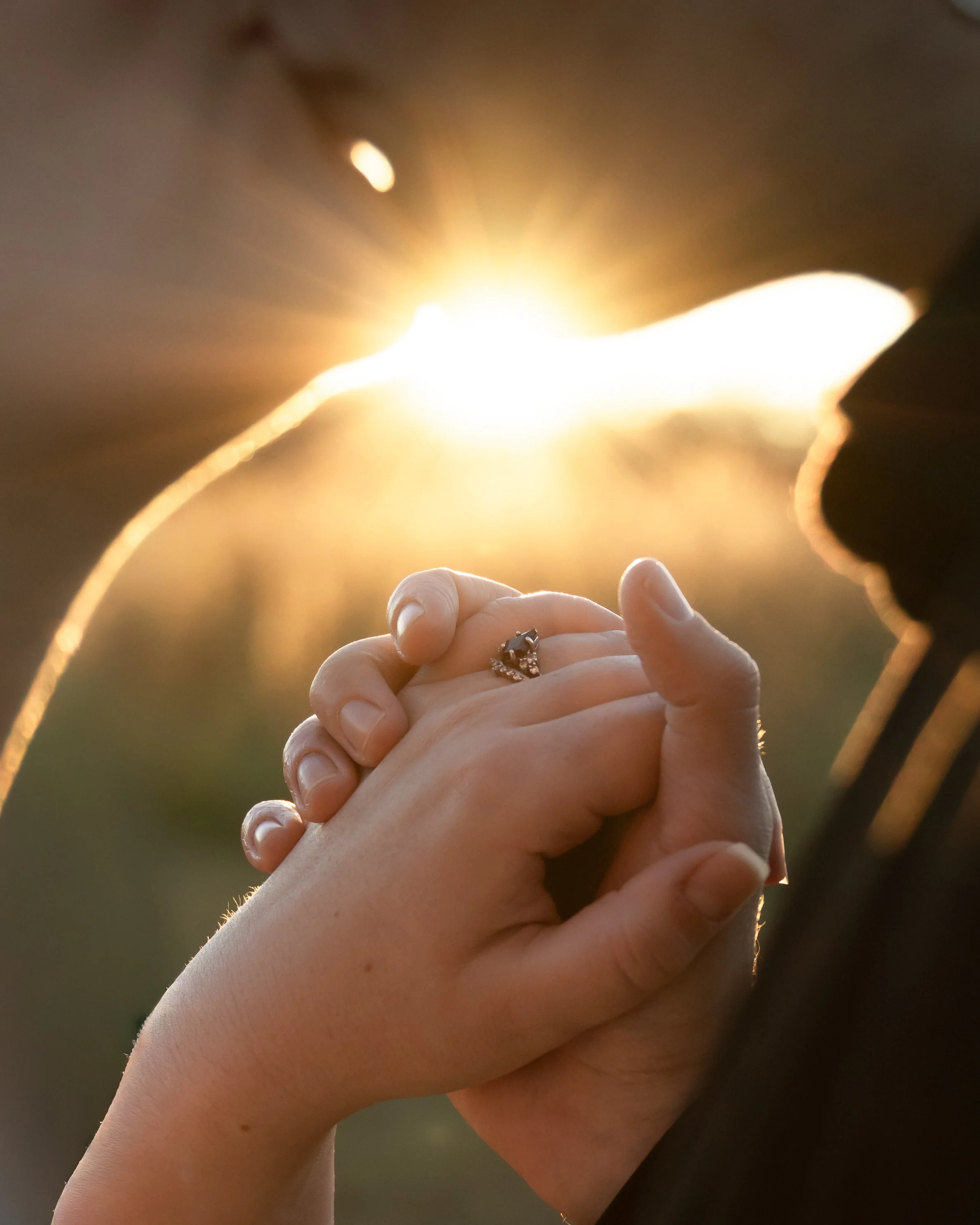 Close-up of a woman and a man holding hands, with the man wearing a wedding ring, sunlight shining brightly in the background.
