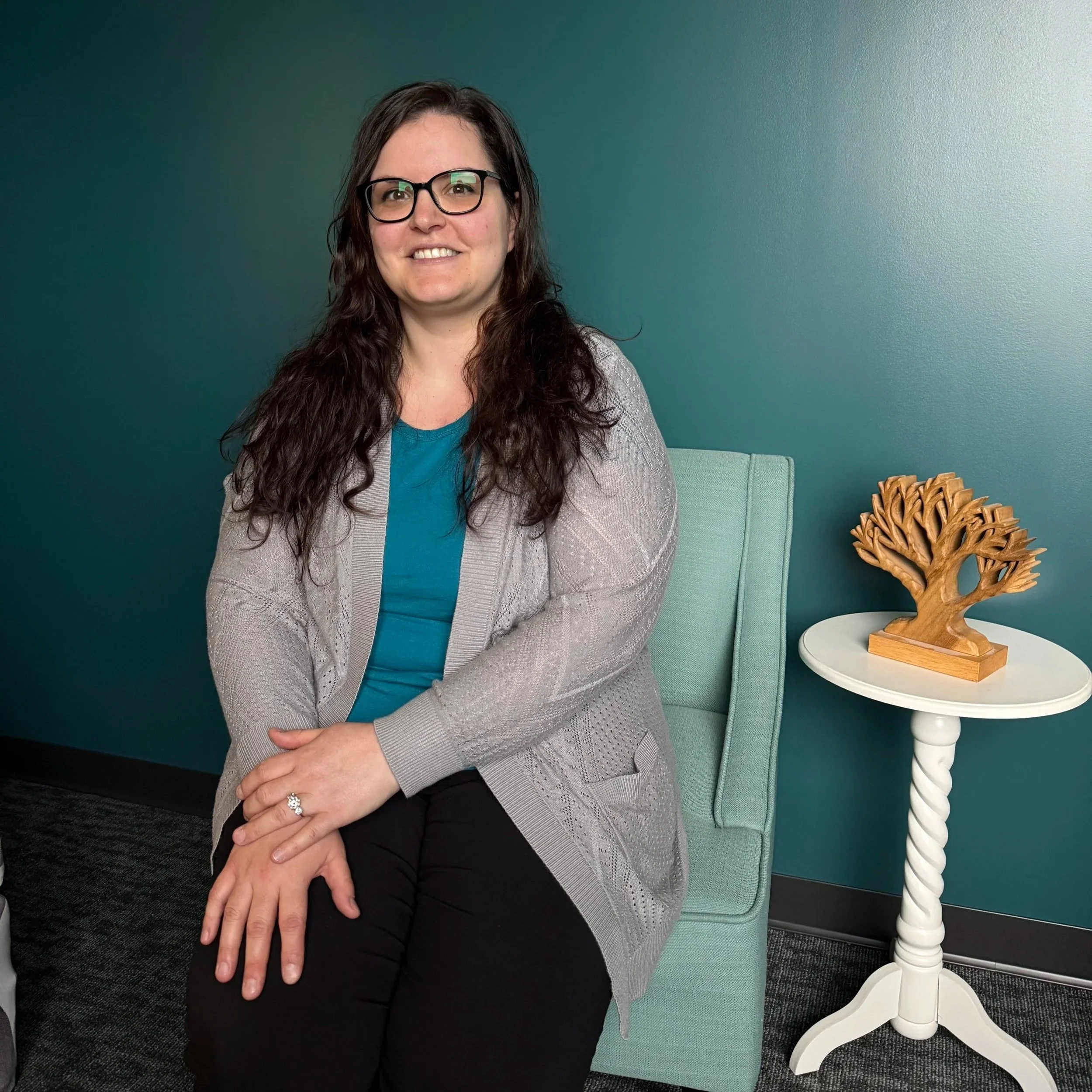A woman with glasses and dark hair, wearing a gray dress, seated indoors near a white wall and a small round table with a decorative white flower plant.