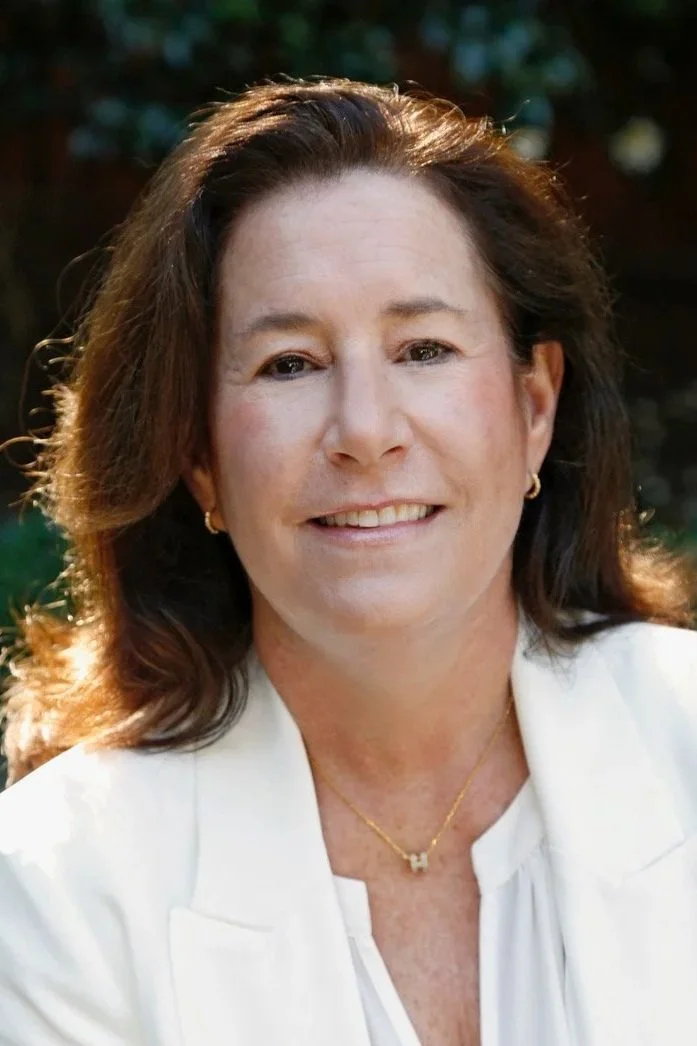 A smiling woman with shoulder-length brown hair, wearing a white blazer, gold earrings, and a gold necklace, outdoors with blurred greenery in the background.