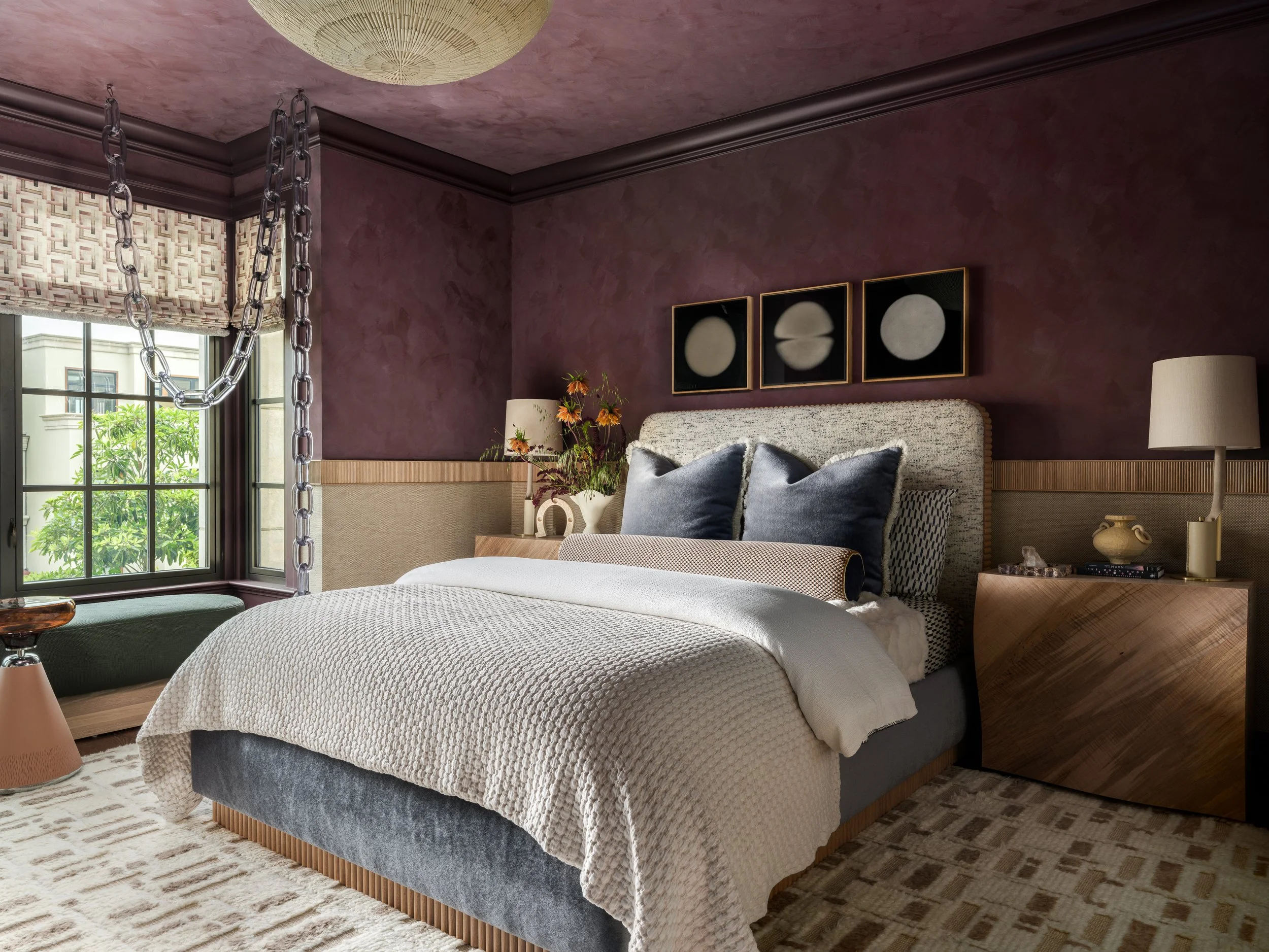 A modern bedroom with purple walls, large bed with textured beige bedding and dark blue pillows, a window with beige blinds, and abstract art depicting lunar phases on the wall.