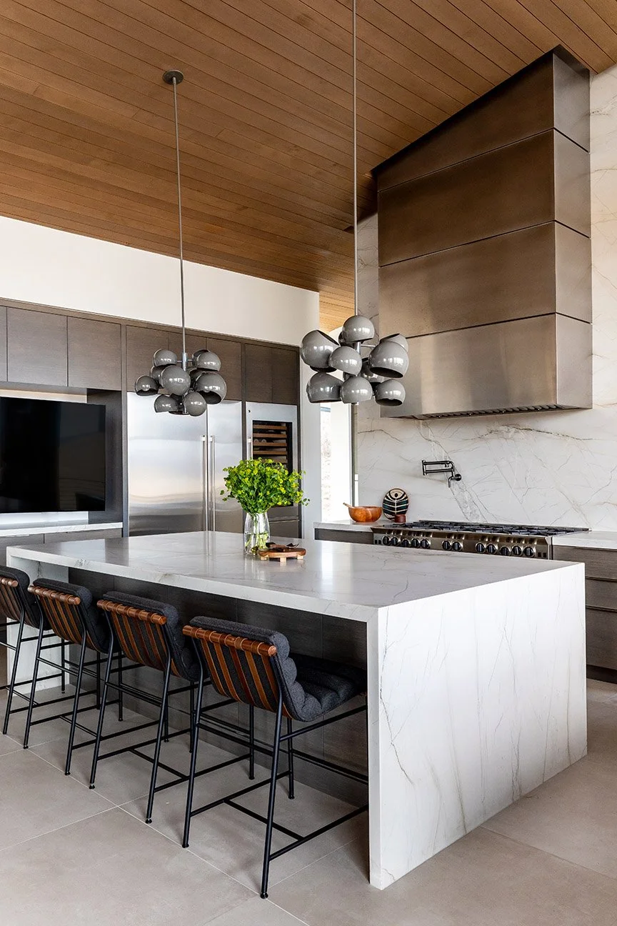 Modern kitchen with a large white marble island, dark wood cabinets, stainless steel appliances, a copper bowl, a potted green plant, and unique gray pendant lights hanging from a wooden ceiling.