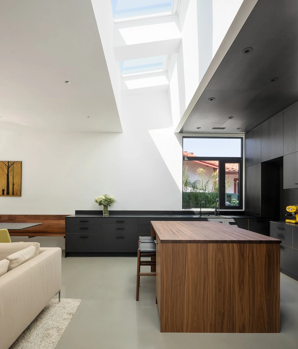 Modern kitchen with black cabinetry, wooden island, large skylight and window showing red-tiled roof and greenery outside.