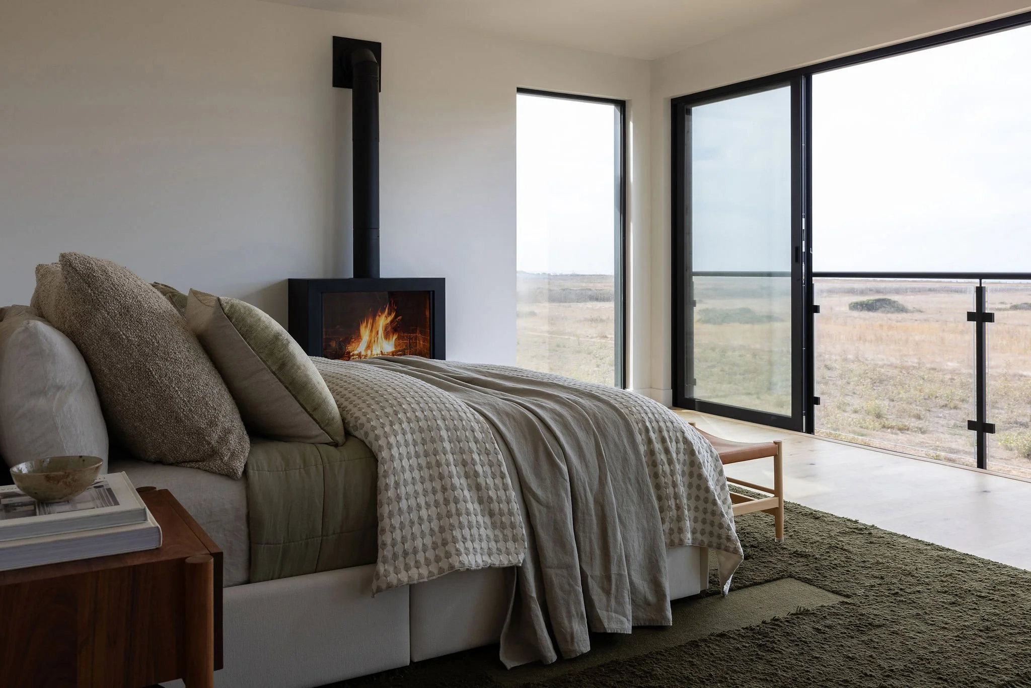 Modern bedroom with a bed, decorative pillows, a blanket, a side table with books and a small bowl, a fireplace with a fire, and large sliding glass doors leading to an outdoor view of a flat landscape.