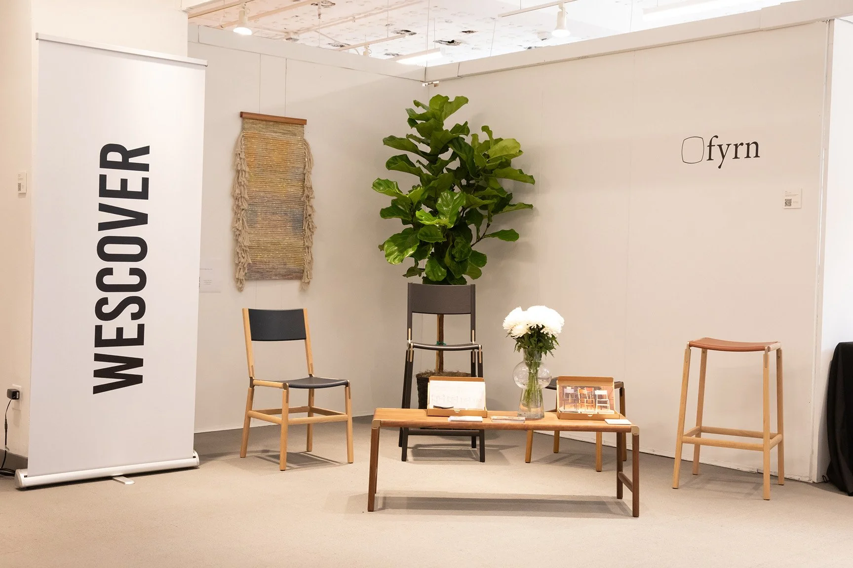 Exhibition booth with a large green plant, a vase of white flowers, a small table, and chairs, with a "WESSCOVER" banner and "fryn" signage on the wall