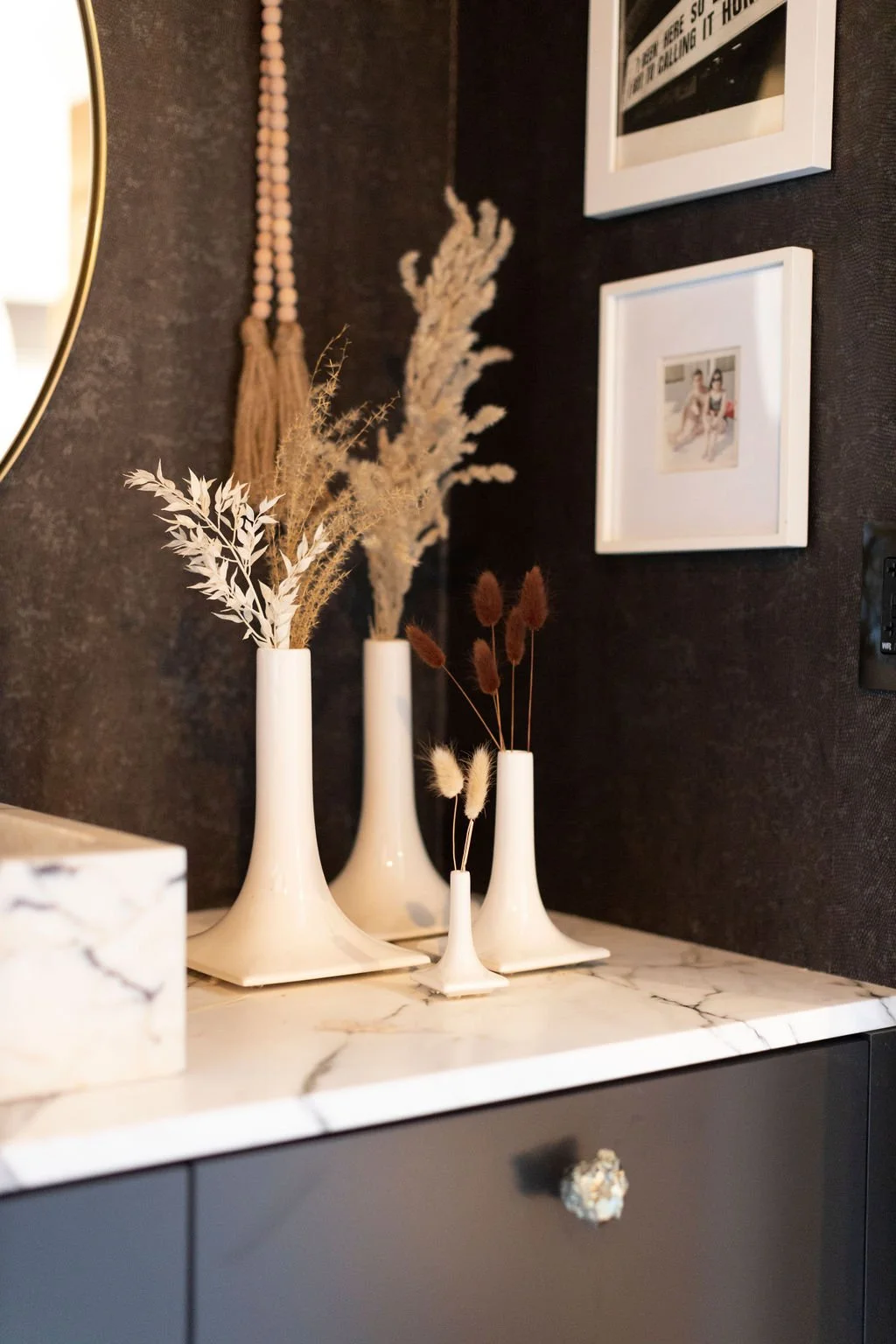 Decorative vases with dried plants on a marble surface, wall art including a beaded tassel and framed pictures in a modern interior.