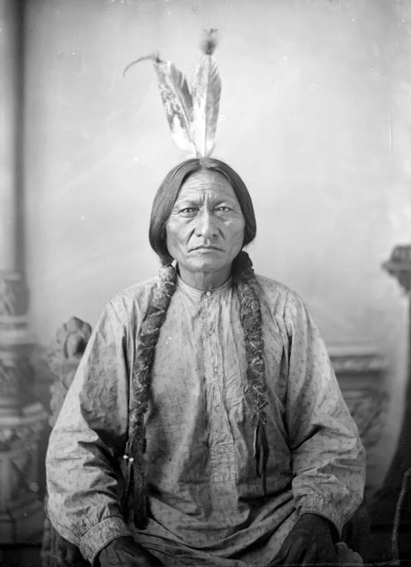 Black and white portrait of a Native American man with braided hair and two feathers in his hair, wearing a patterned shirt.