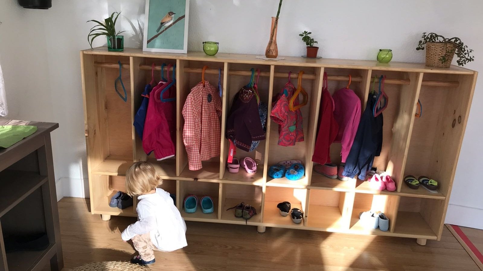 Niño pequeño agachado frente a un estante de madera con abrigos y zapatos, varias perchas de colores, y plantas en la parte superior.