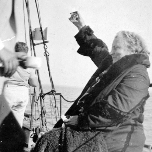 Maria Montessori en un barco levantando un vaso, cielo despejado.