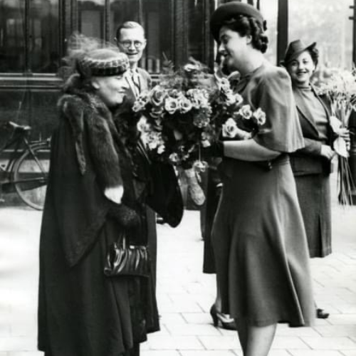 Grupo de personas en la calle, Maria Montessori, una mujer mayor recibe un ramo de flores de otra mujer. Están bien vestidas, en lo que parece ser una época anterior. Hay bicicletas en el fondo.