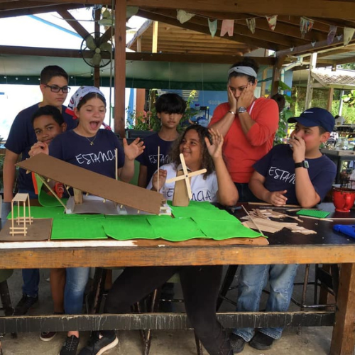Un grupo de jóvenes y un adulto con maquetas de proyectos escolares en una mesa al aire libre.