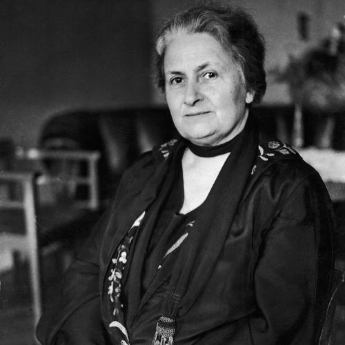 Maria Montessori, una mujer mayor sentada en un sillón en un entorno interior, vistiendo ropa formal y mirando hacia la cámara.