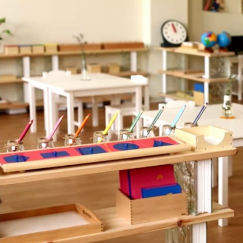Aula de escuela Montessori con mesas blancas y estanterías de madera, elementos educativos como lápices de colores y materiales de aprendizaje.