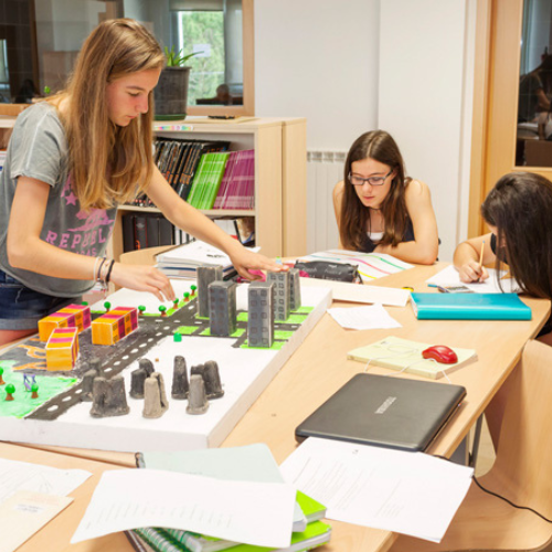 Tres estudiantes trabajando en un proyecto escolar, con una maqueta de una ciudad sobre la mesa, rodeados de libros y cuadernos, en un aula.