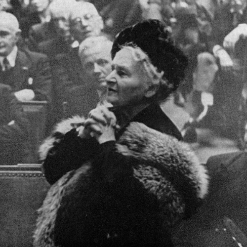 Maria Montessori, mujer mayor vestida con abrigo de piel en un evento formal, rodeada de personas, posiblemente en una reunión o conferencia.