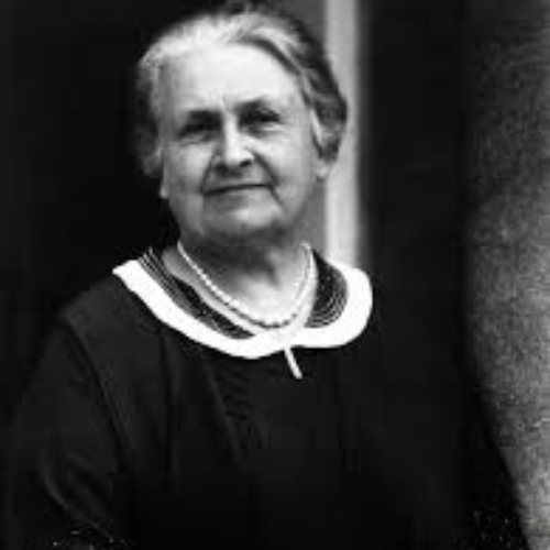 Maria Montessori, una mujer mayor con un collar de perlas, vestida de manera formal, mira a la cámara en una fotografía en blanco y negro.