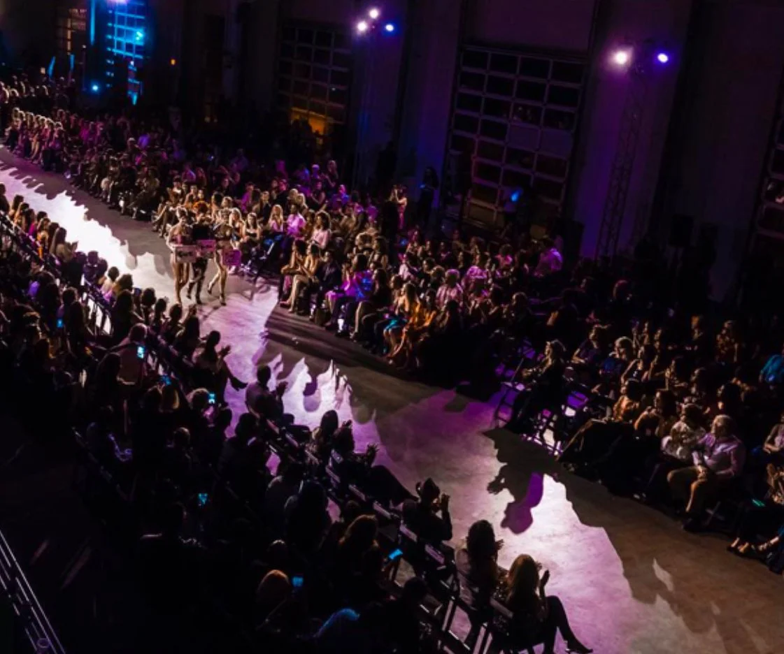 Audience seated in a darkened auditorium watching a fashion show with models walking the runway, illuminated by purple and blue lighting.