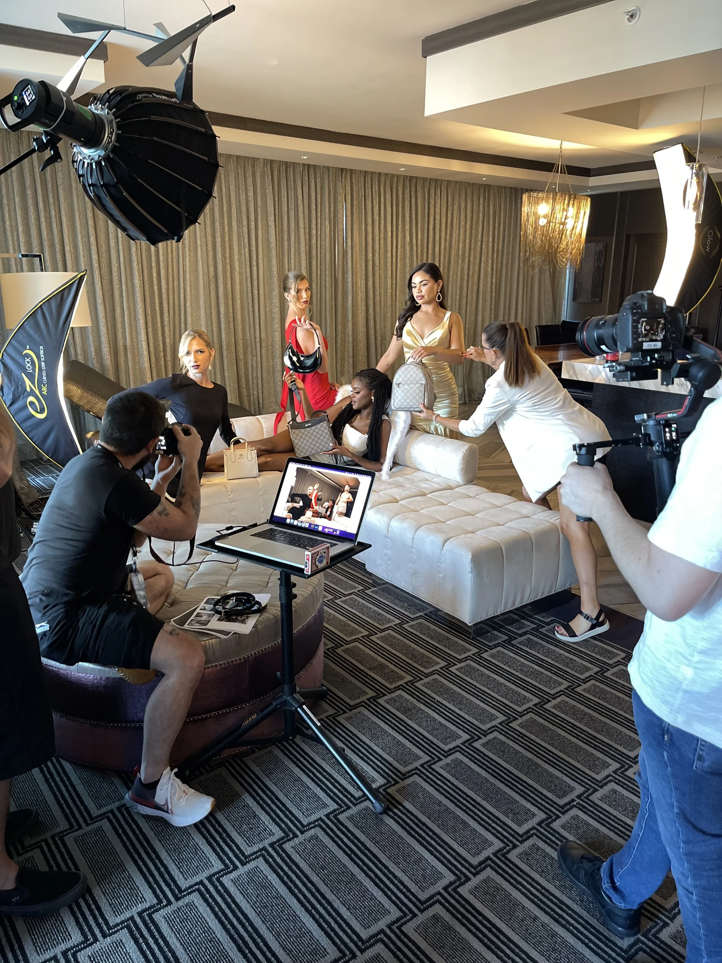 A photoshoot with five women and six crew members in a hotel room, featuring studio lighting and cameras, with three women sitting on a sofa and two women standing, one holding a purse and another holding a bag.
