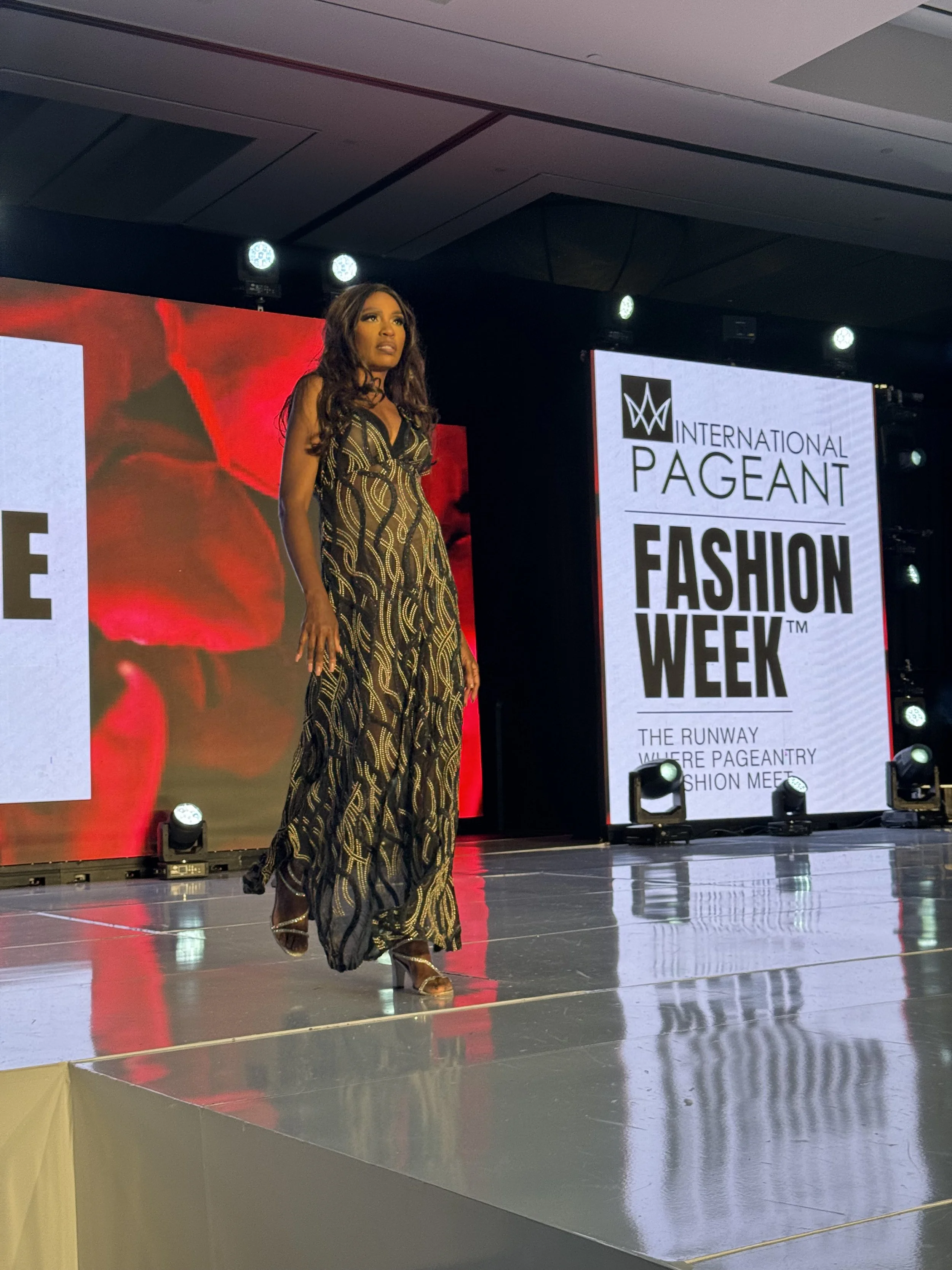 A woman walking on runway during Fashion Week at an international pageant event, wearing a long, black and gold patterned dress and high heels, with a large digital screen behind her displaying event signage.