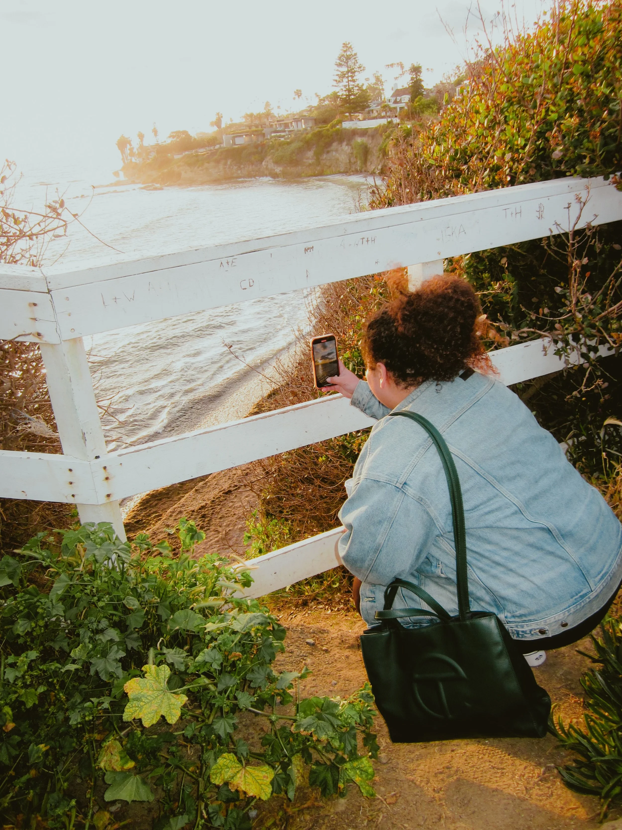 A woman with curly hair wearing a denim jacket squatting down near a white wooden fence, taking a photo with her phone of a coastal scene with houses, trees, and water during sunset.