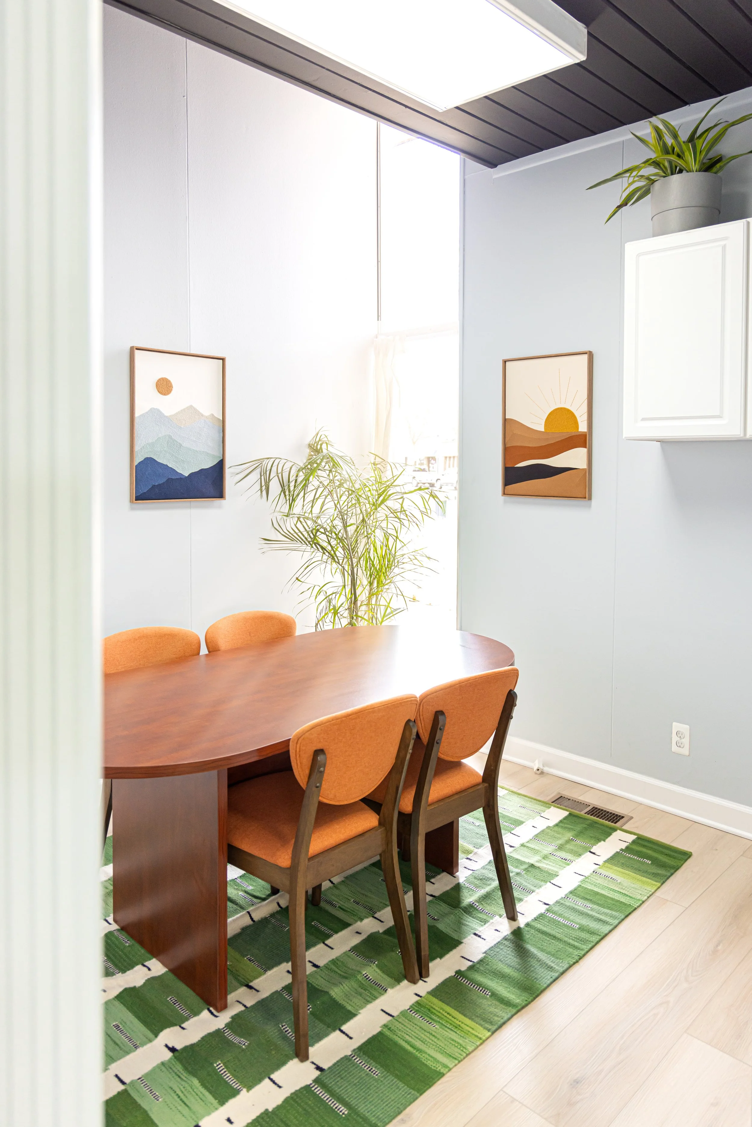 A conference room with a wooden table, six orange cushioned chairs, framed artwork of mountain landscapes on the walls, a green patterned rug, and a potted plant near a large window.