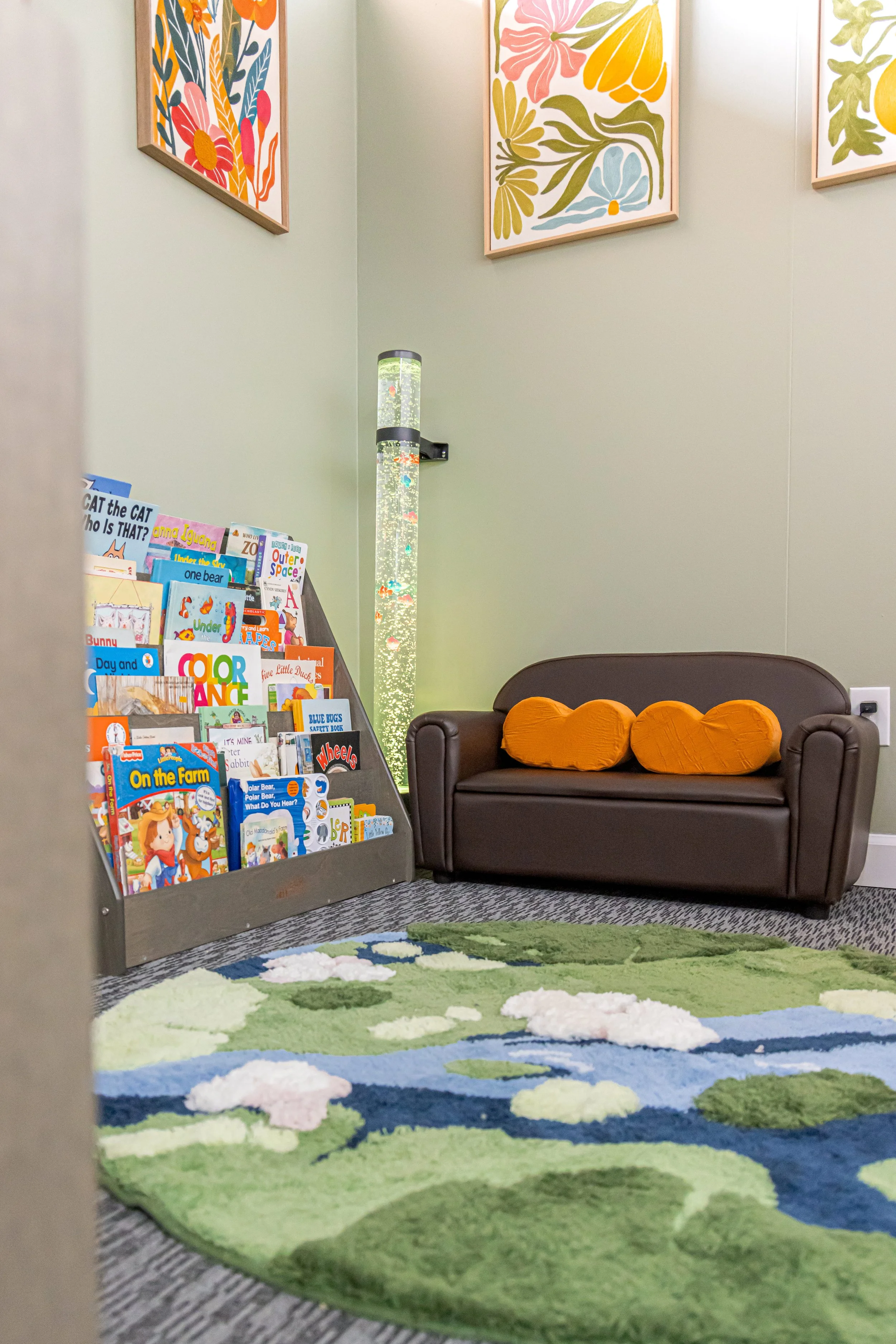 A waiting area with a colorful floral-themed rug, a small book display with children's books, a dark brown sofa with orange cushions, a bubble tube light fixture on the wall, and framed floral artwork on the green wall.