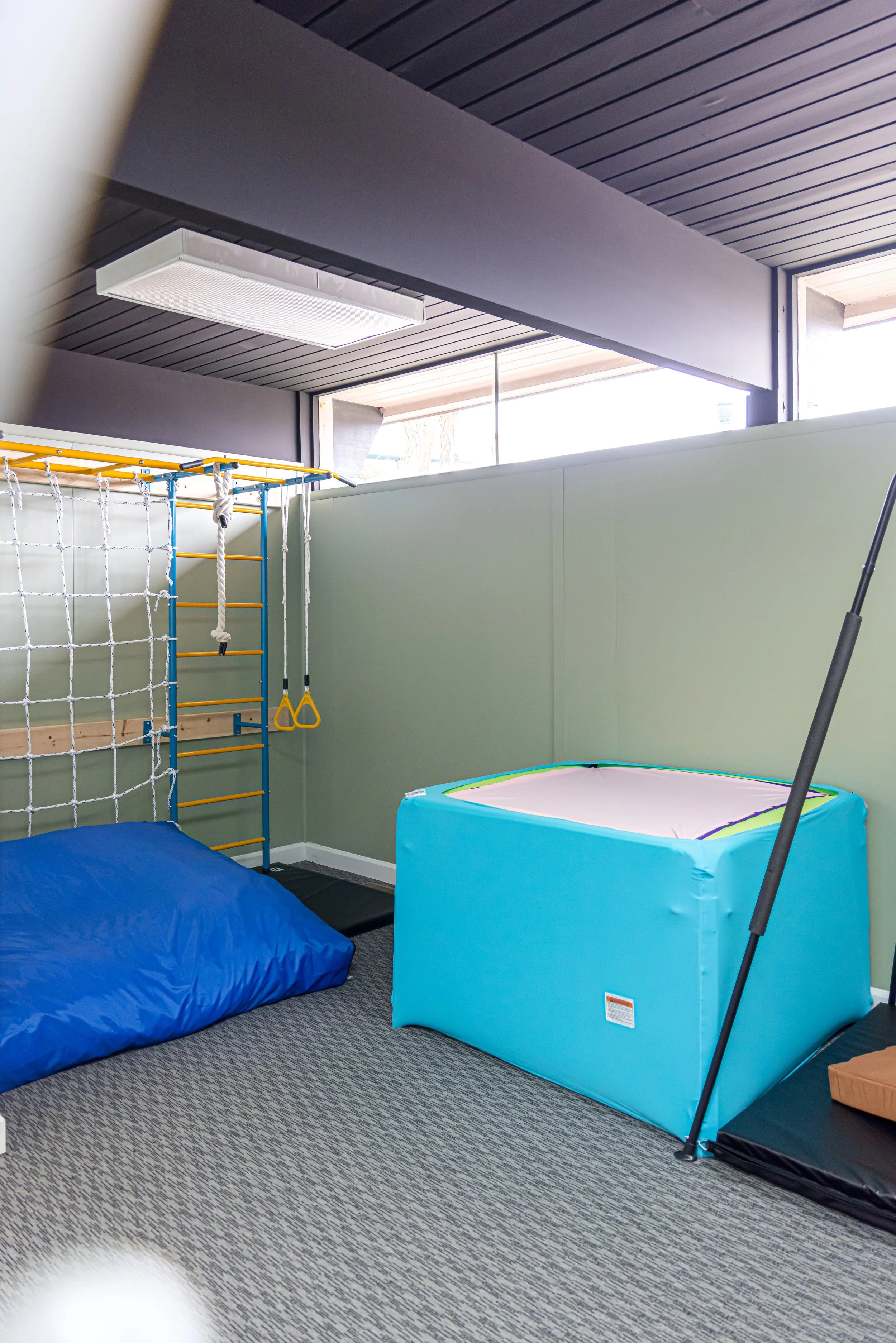 Indoor play area with soft mats, climbing ropes, and a small trampoline on a carpeted floor, in a room with large windows and dark ceiling panels.
