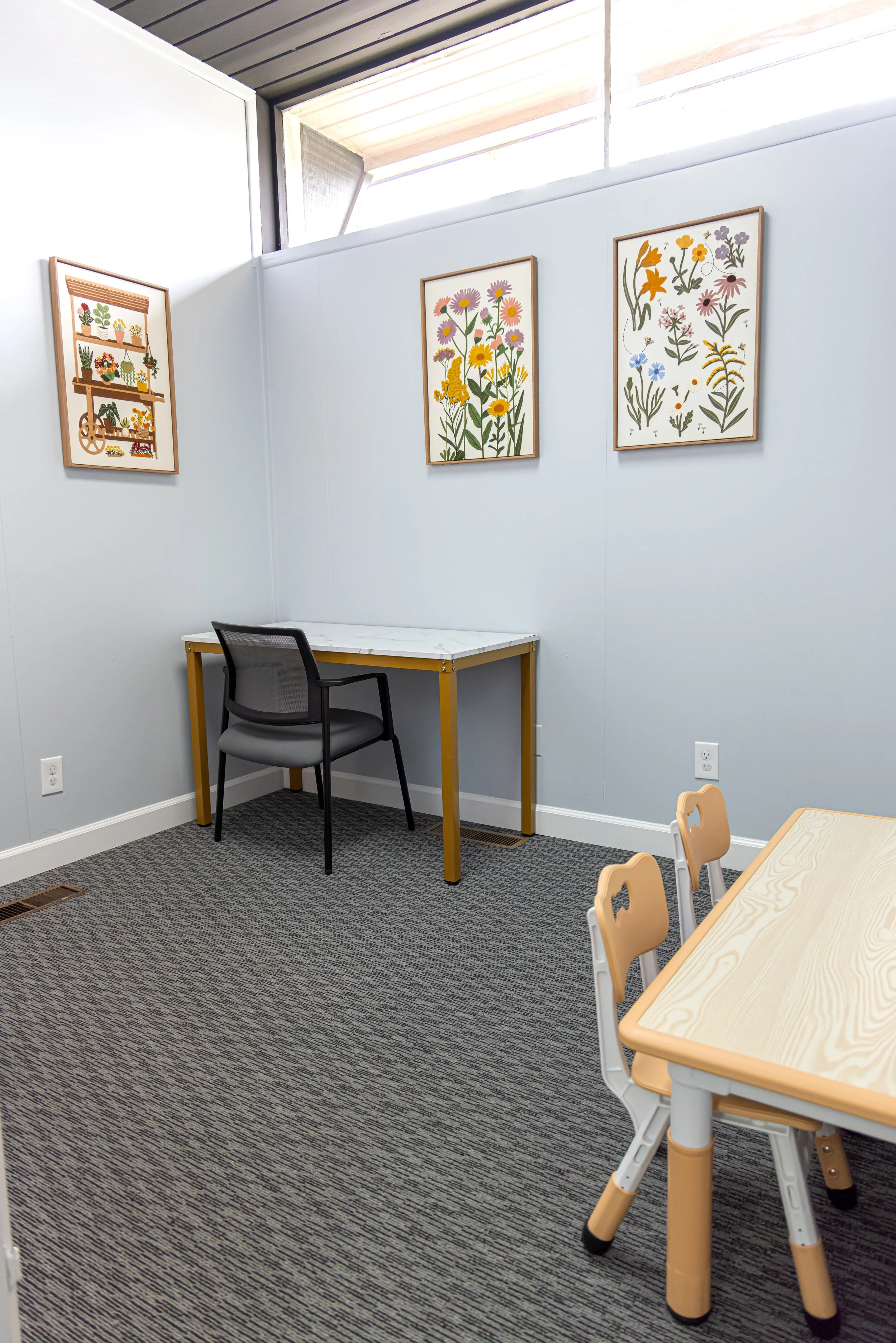 A small room with a high ceiling and white walls. There is a yellow desk with a black and gray office chair, and a wooden high chair with white legs near a rectangular light wood table with rounded edges. On the wall, there are three framed botanical illustrations of various flowers, and there are windows allowing natural light to enter.