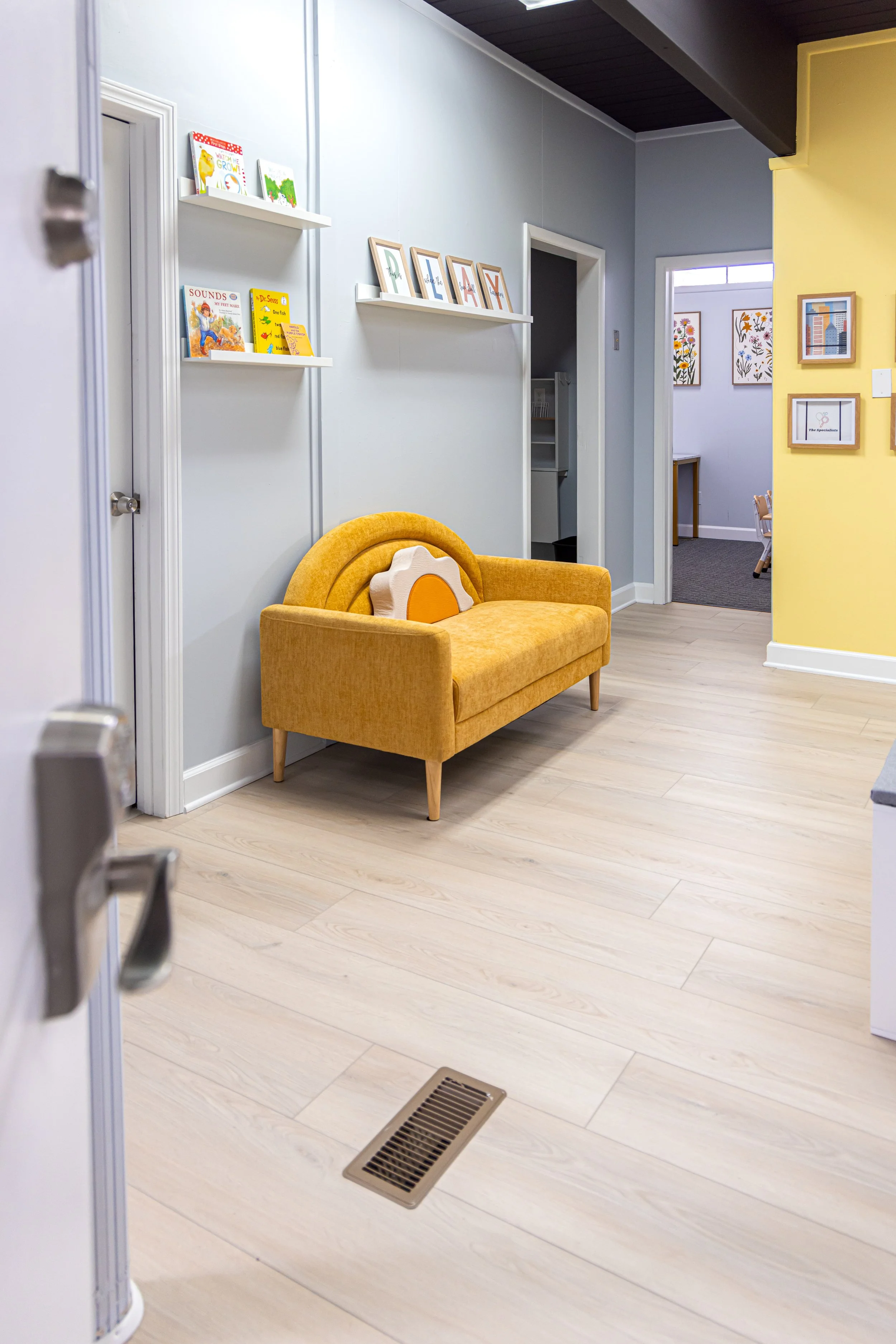 Interior of a room with a yellow sofa, light-colored wooden floor, and white walls decorated with children's books and framed artwork. Part of a door and floor vent are visible.