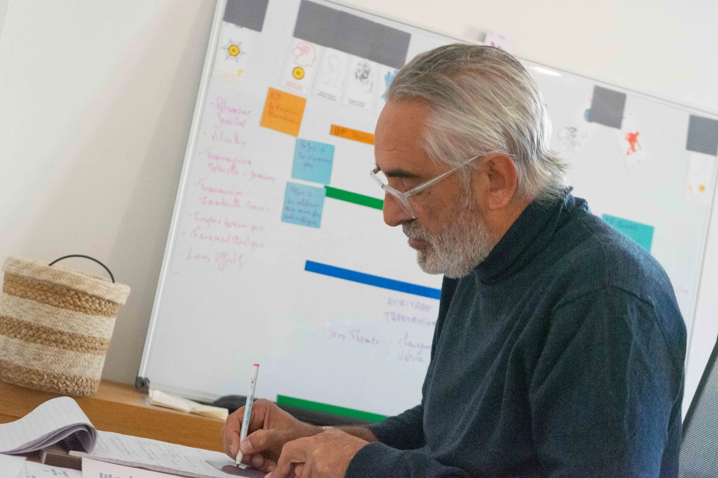 Un homme âgé avec des cheveux gris et une barbe, portant des lunettes, qui écrit dans un cahier. En arrière-plan, un tableau blanc avec des notes autocollantes colorées et des écrits.