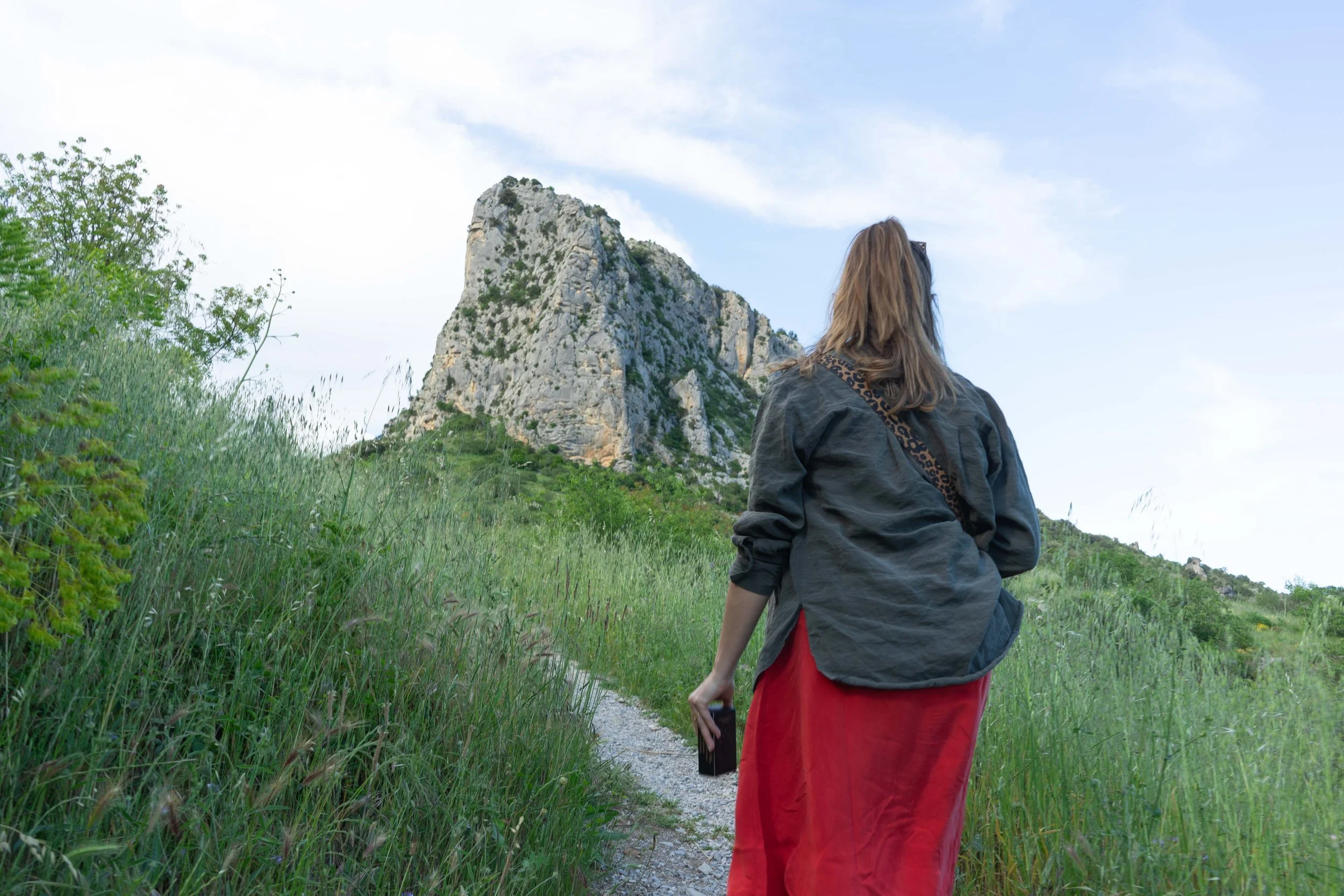 Une femme marche sur un sentier entouré d'herbes vertes, avec une montagne rocheuse en arrière-plan, sous un ciel partiellement nuageux.