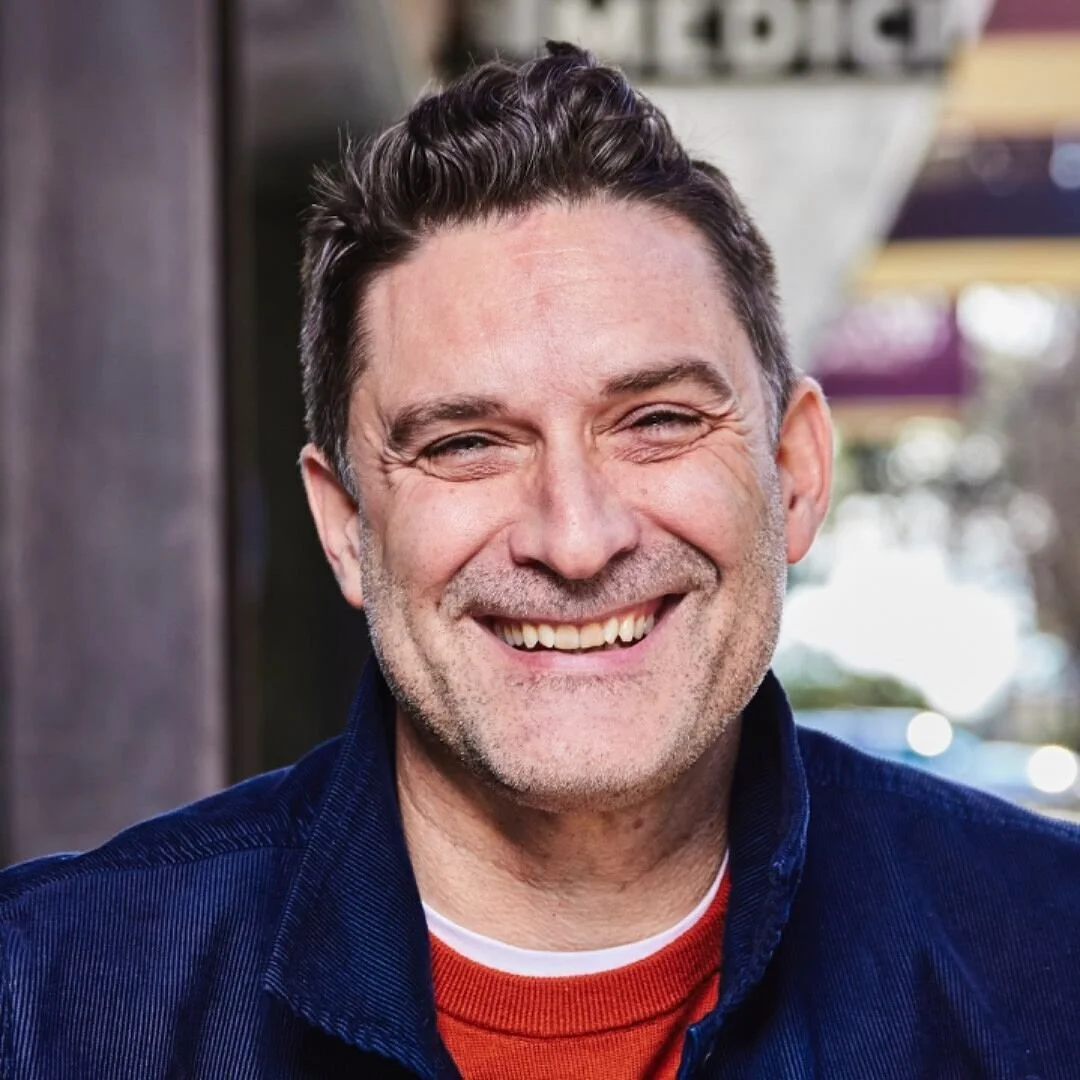 Close-up of a smiling middle-aged man with dark hair and a beard, wearing a blue jacket and red shirt, outdoors with blurred background.