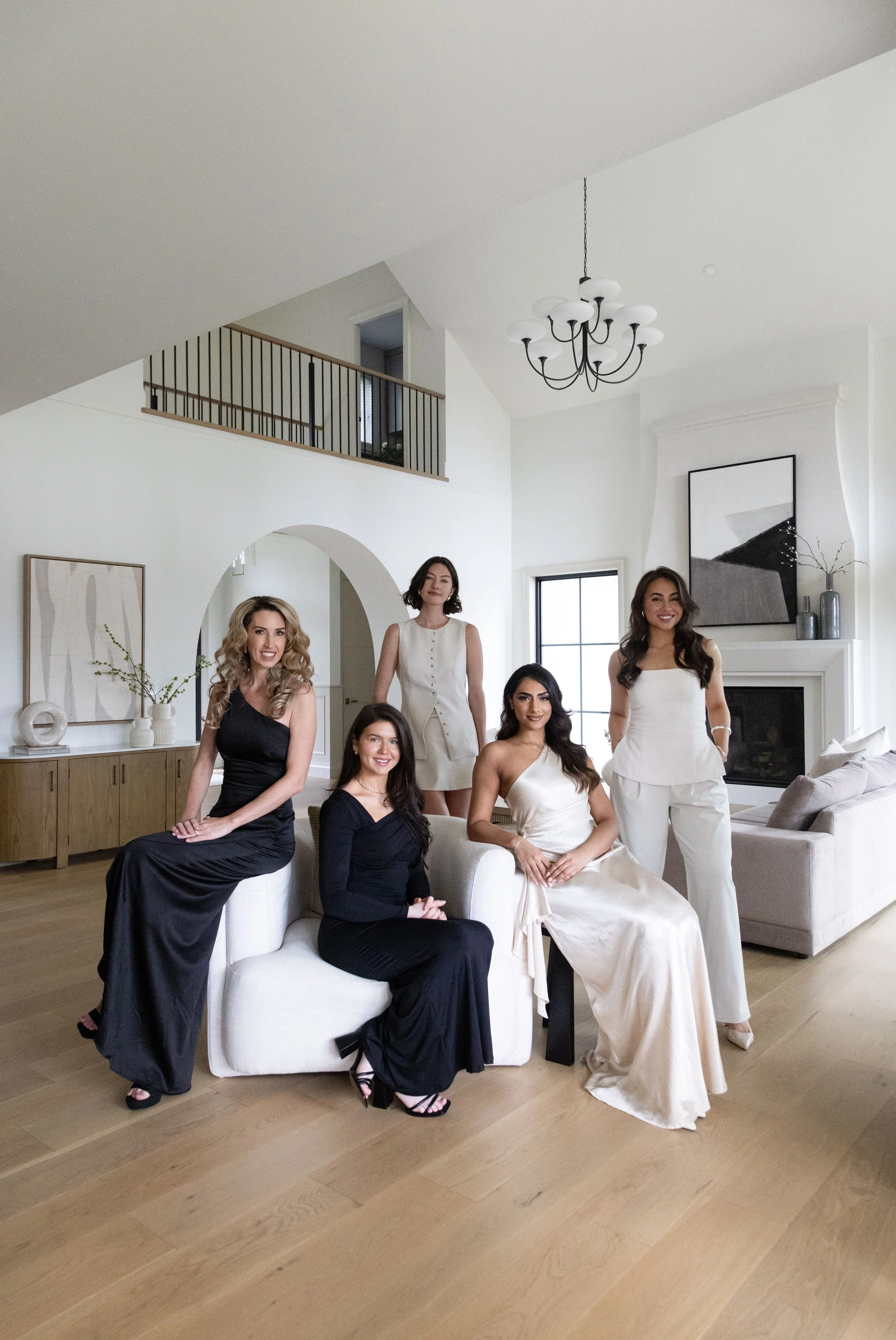 Five women in elegant dresses sitting and standing in a modern, minimalist living room with white walls, wooden floors, and large windows.