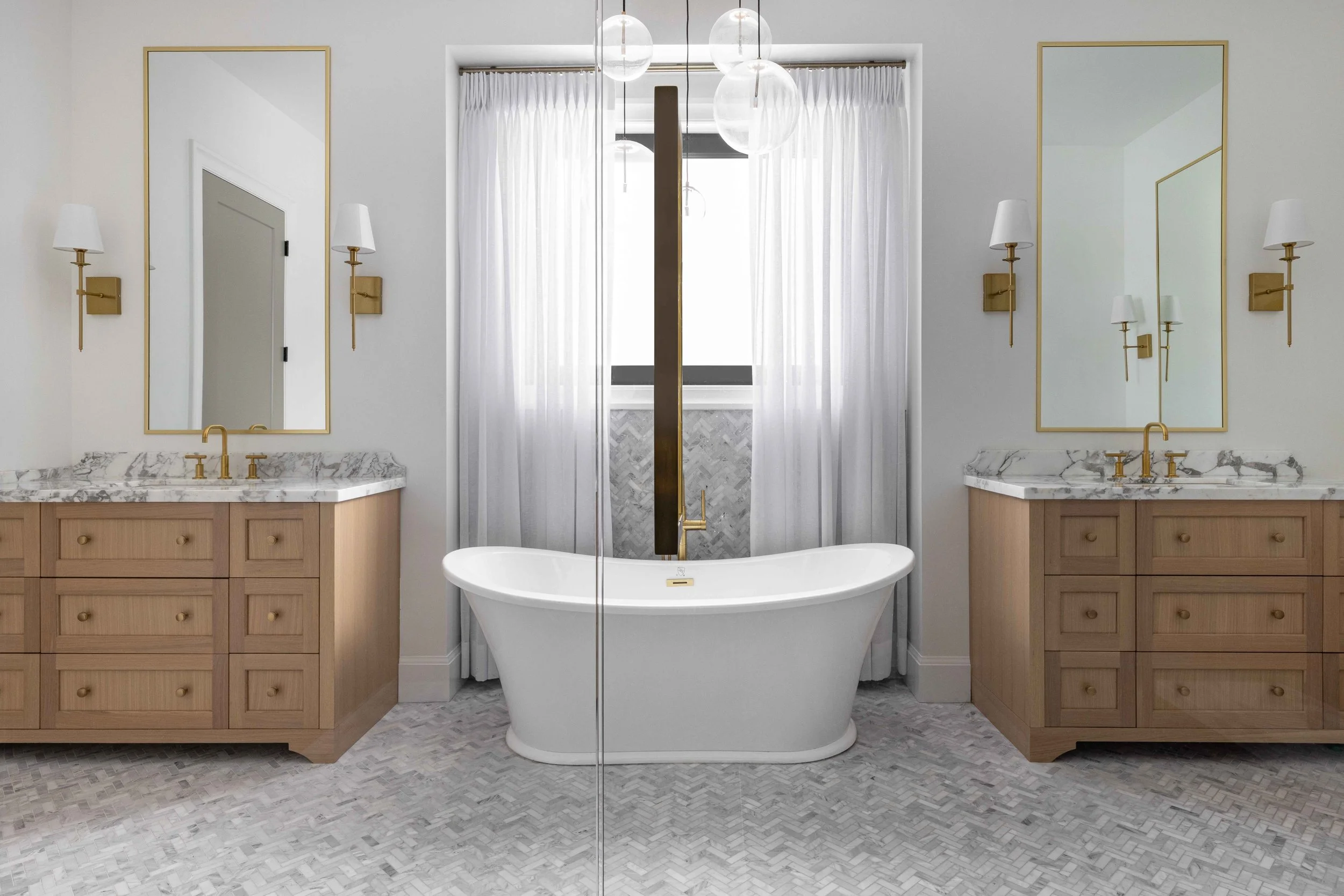 Symmetrical bathroom with a free-standing bathtub in the middle, flanked by two wooden vanities with marble countertops and gold fixtures. Behind the tub, there is a window with sheer white curtains and modern hanging light fixtures. Large mirrors with gold frames are mounted above each vanity, paired with wall sconces.
