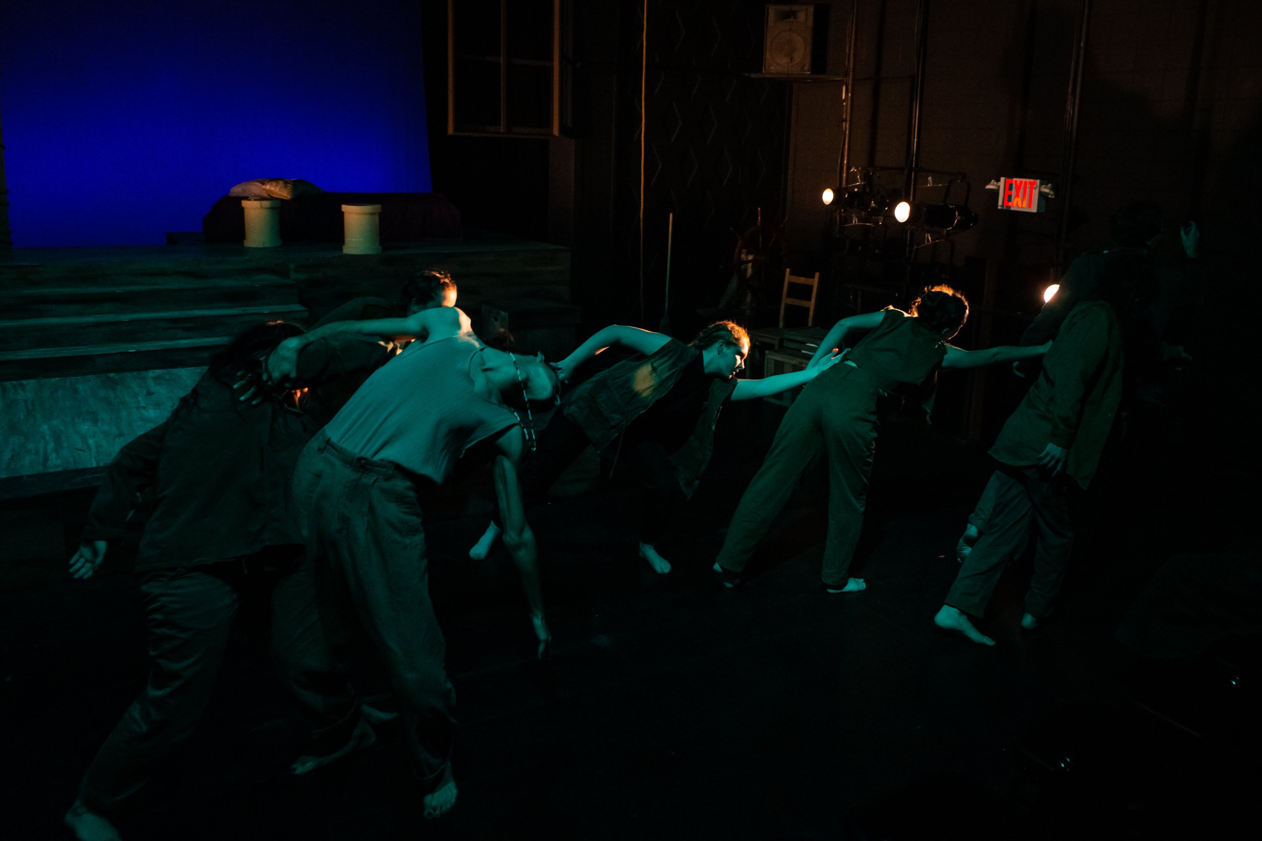 Performers on stage in dim lighting with a blue backdrop, dressed in casual attire, engaged in expressive movement, possibly part of a theatrical or dance performance.
