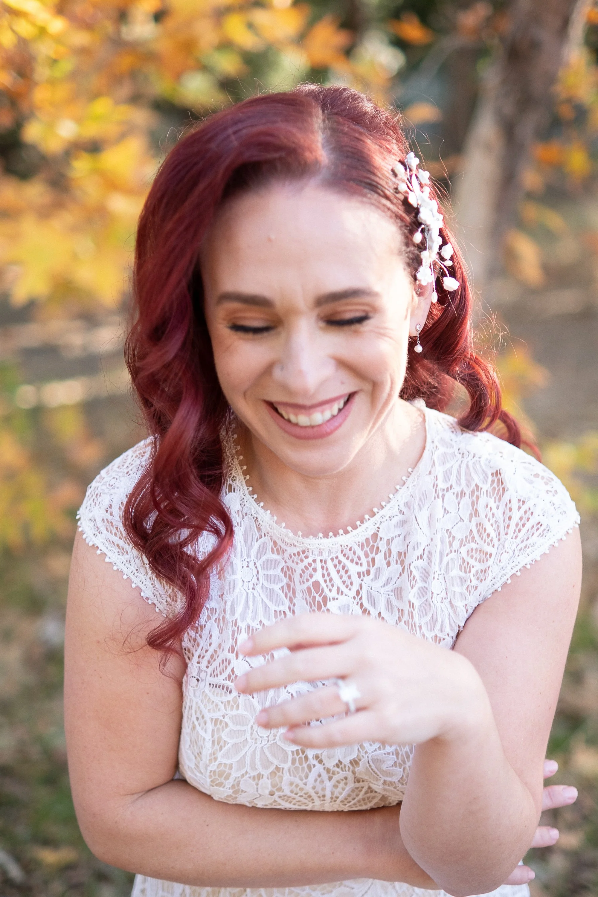 A smiling woman with red hair and a white lace dress is outdoors in a fall setting. She has floral hair accessories and is looking down, appearing happy and joyful.