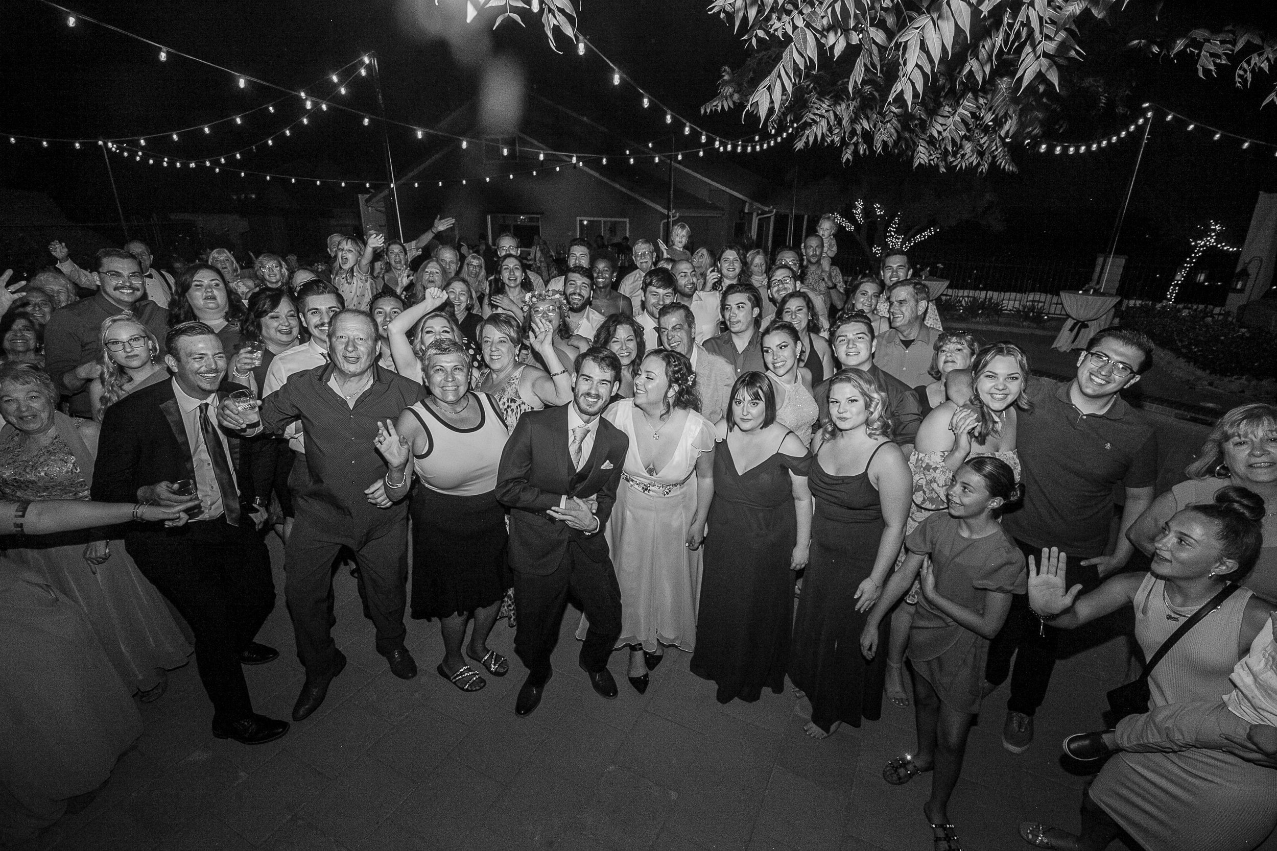 Black and white photo of a large group of people celebrating at an outdoor event under string lights at night, with some smiling and gesturing.