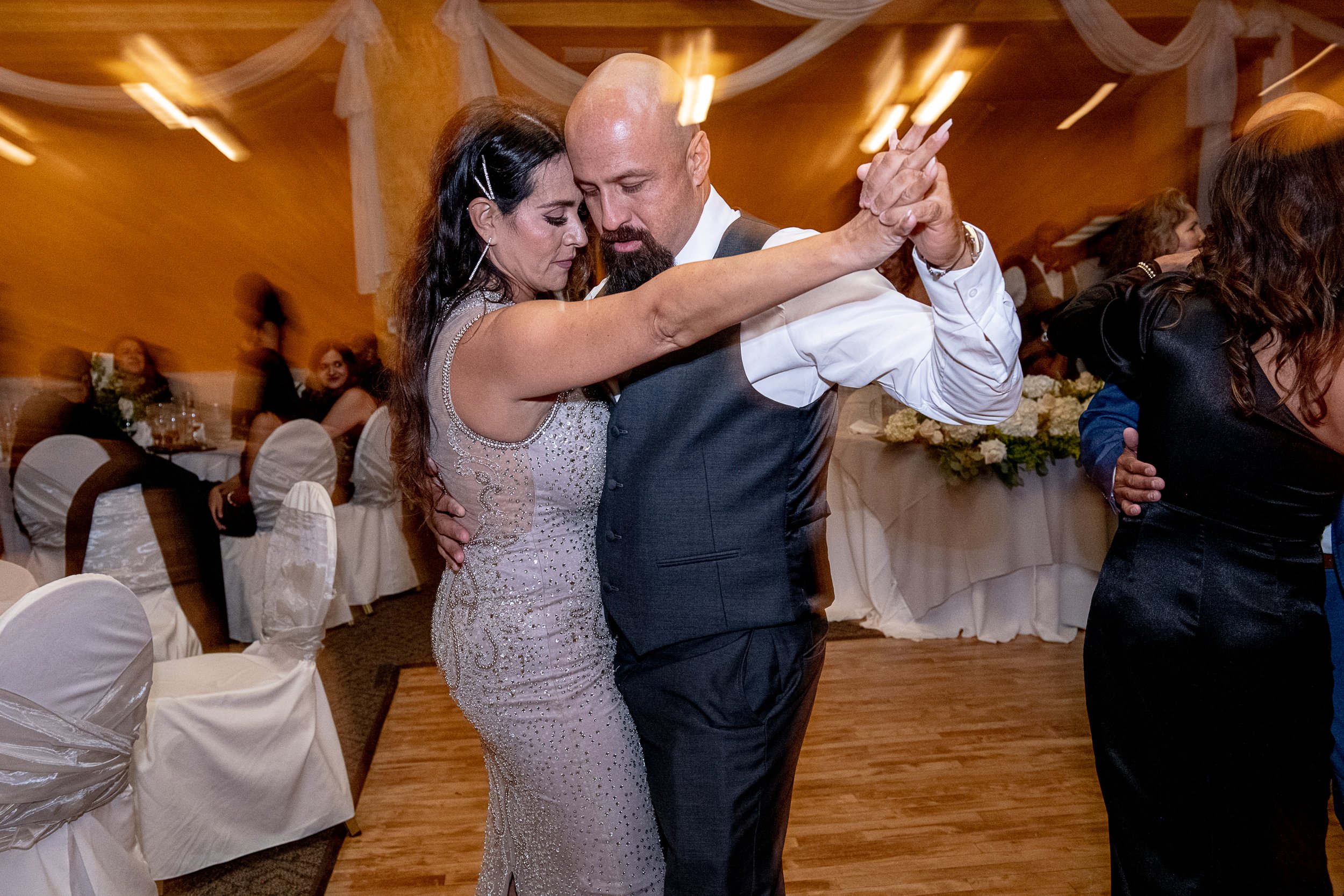 A couple dancing closely at a wedding reception, with the woman wearing a beaded gown and the man in a vest and dress shirt, surrounded by seated guests and draped decor.